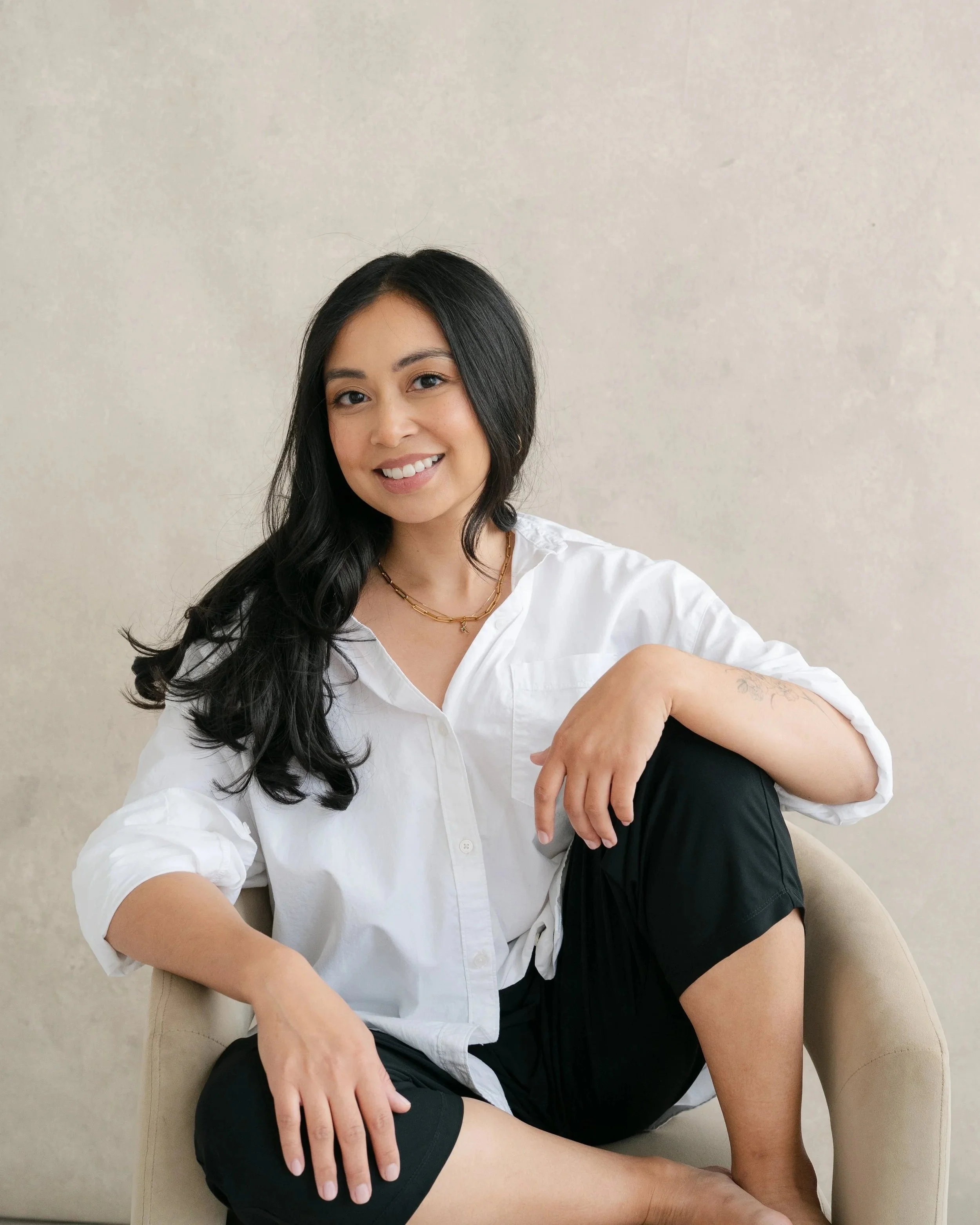 A woman with long black hair, wearing a white shirt and black pants, sitting barefoot on a beige chair against a plain light-colored wall, smiling at the camera.
