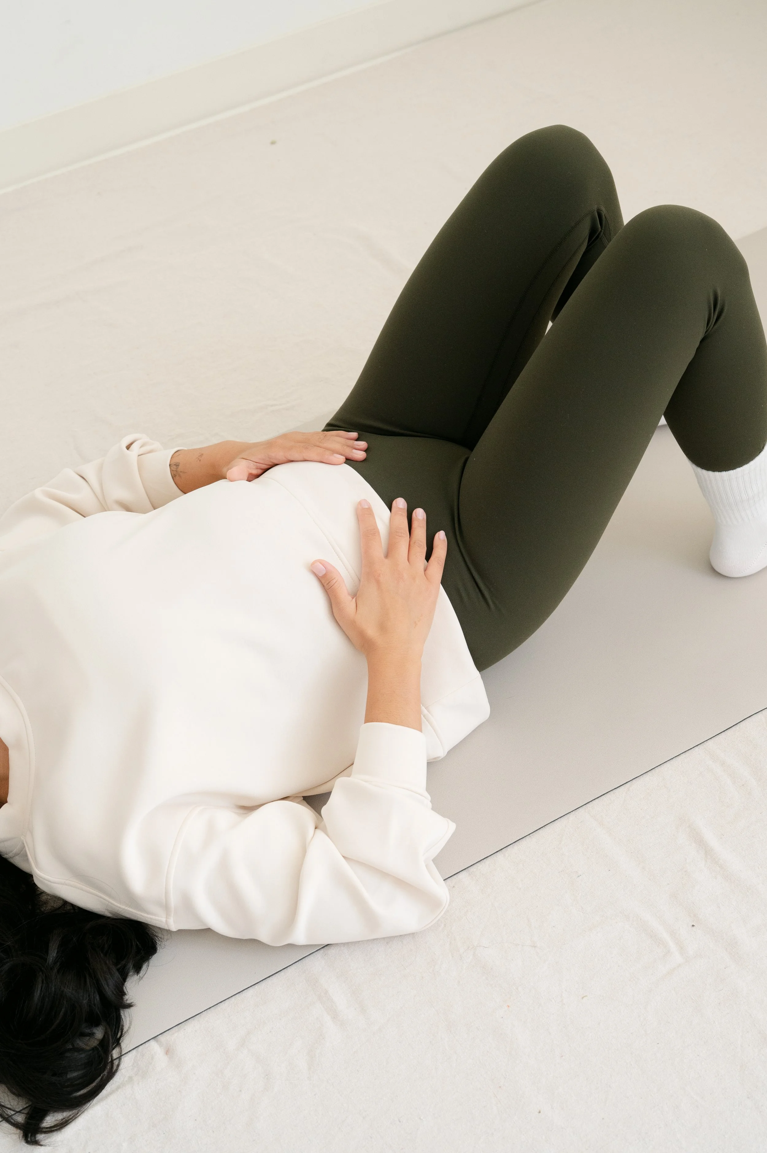 A person lying on their back on a yoga mat, with hands resting on their lower abdomen, legs bent at the knees, in a yoga or relaxation pose.