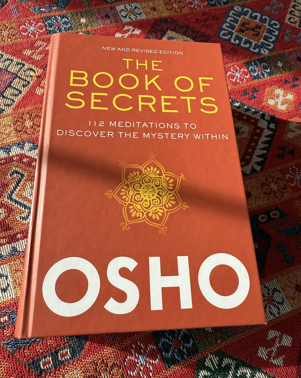 My copy of Osho's Book of Secrets, laid out on my own tablecloth.  It's a big fat red book, and the name of the author O S H O is spelled out in identical huge text on the bottom of the cover just like the corrner of the book we see on Epstein's desk