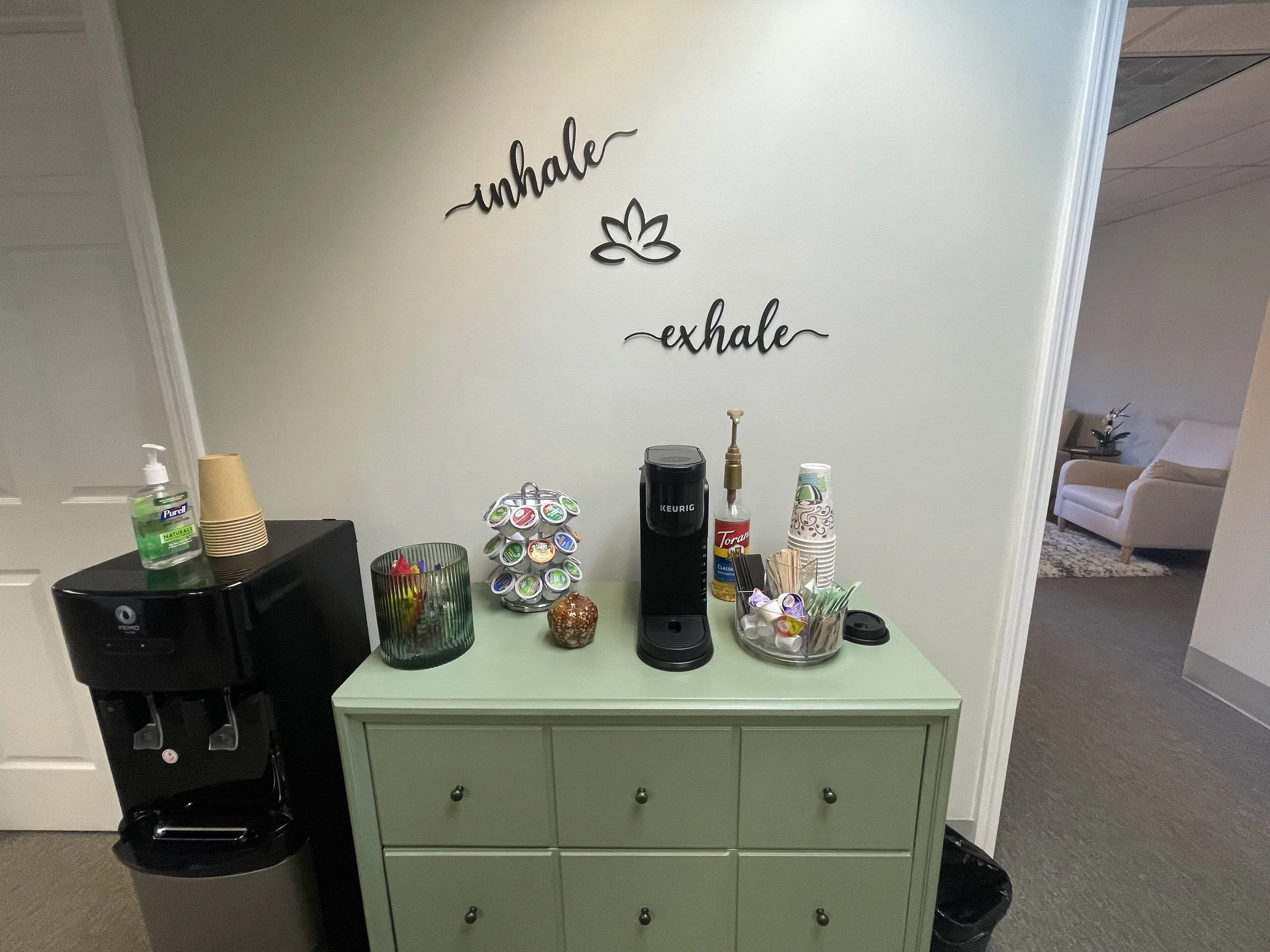 Living room with a gray sofa, patterned pillows, a wooden desk with a laptop, potted plants, a beige shag rug, a gray armchair, a side table with a lamp, and two windows with blinds.