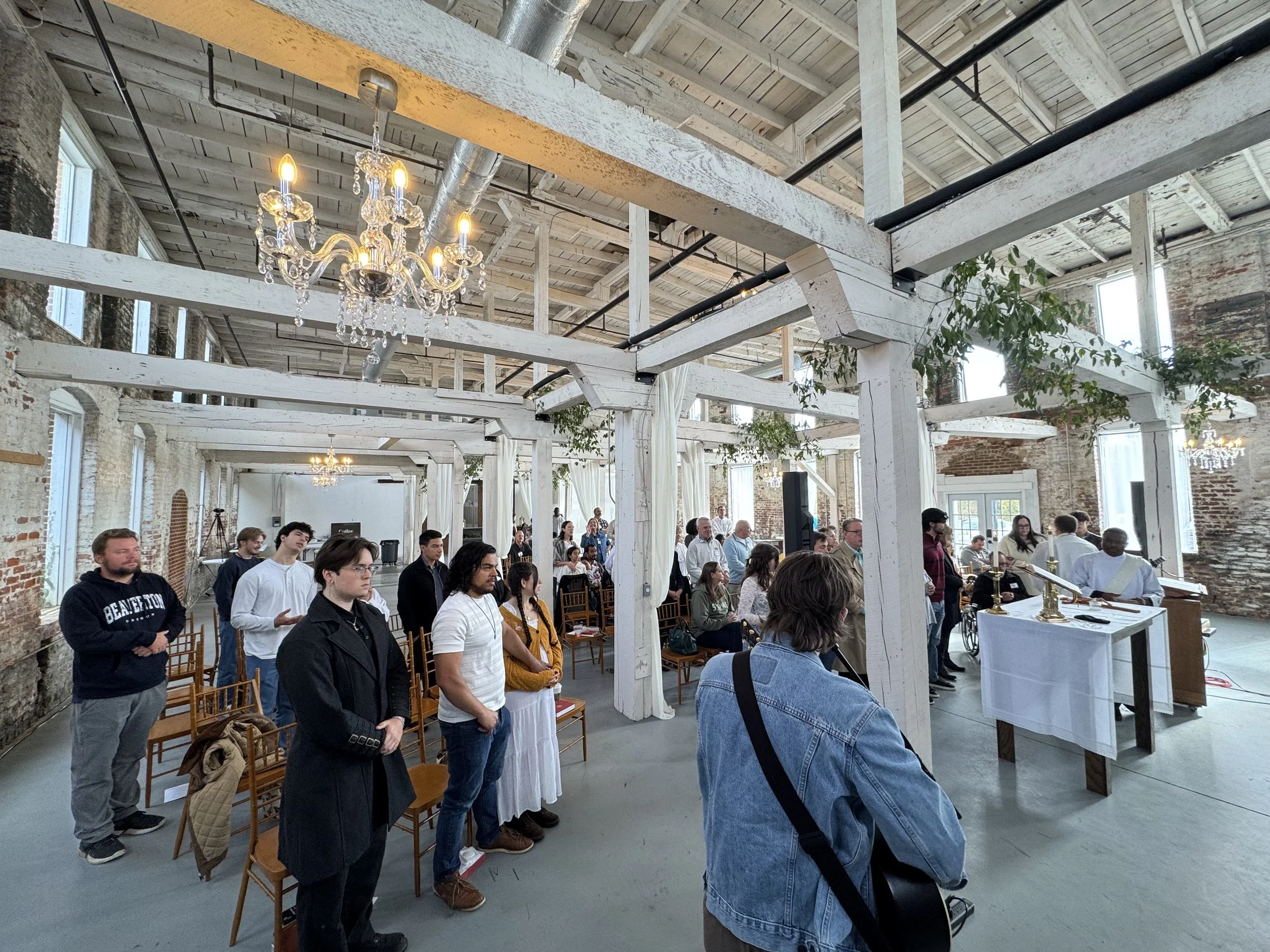 People gathered inside a spacious industrial-style venue with exposed brick walls, white wooden beams, and chandeliers, participating in a ceremony or event.