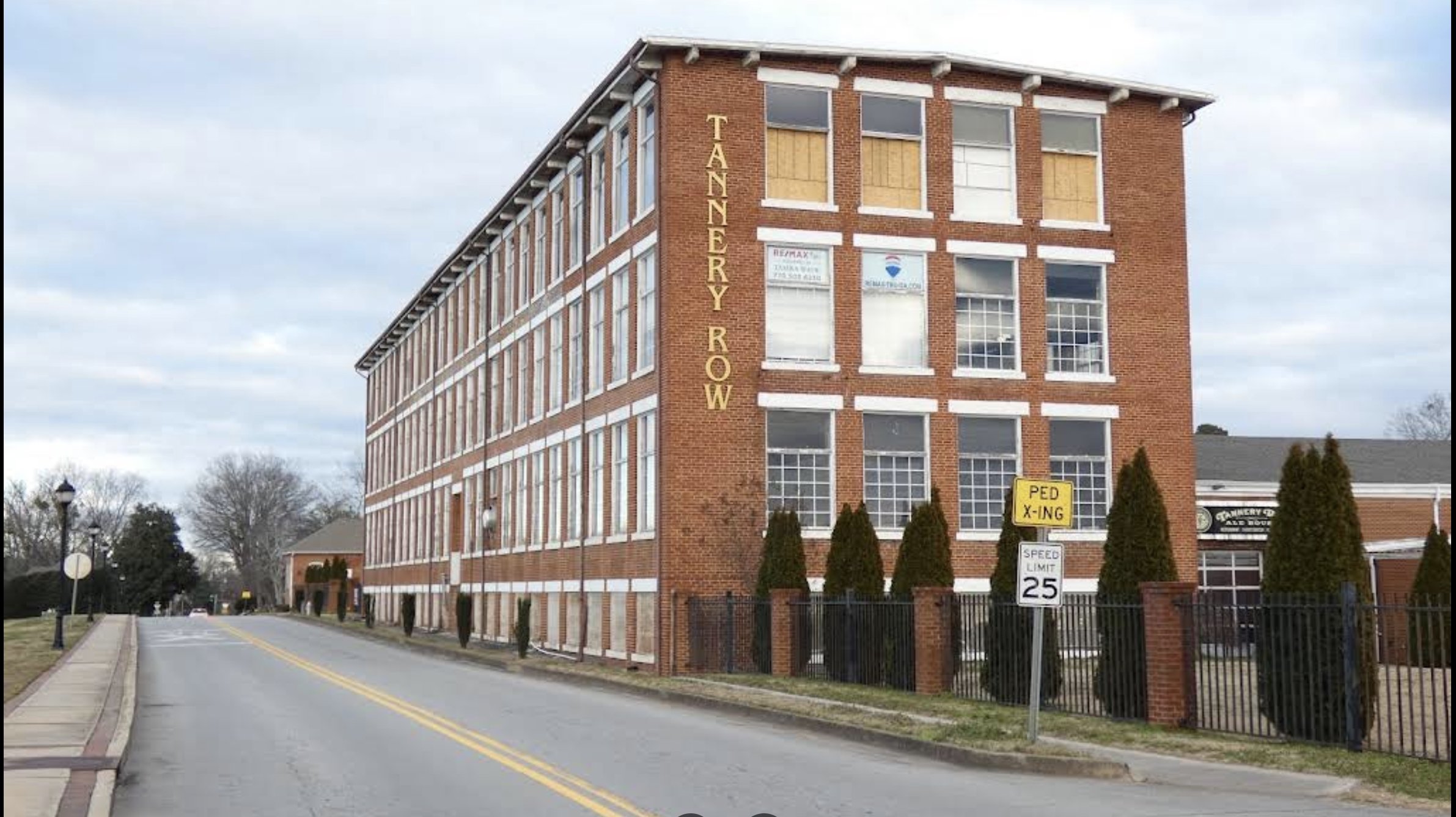 A four-story red brick building with large windows, labeled 'TANNERY ROW' on the side, located on a quiet street with a sidewalk, trees, and a fence in front.
