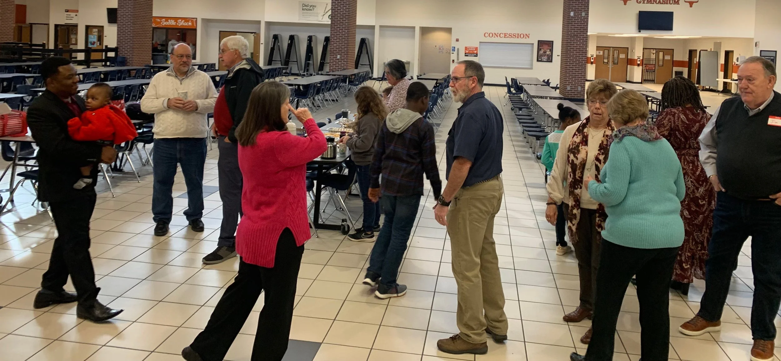 Group of people gathering and talking in a school cafeteria with tables, chairs, and concession stand in the background.