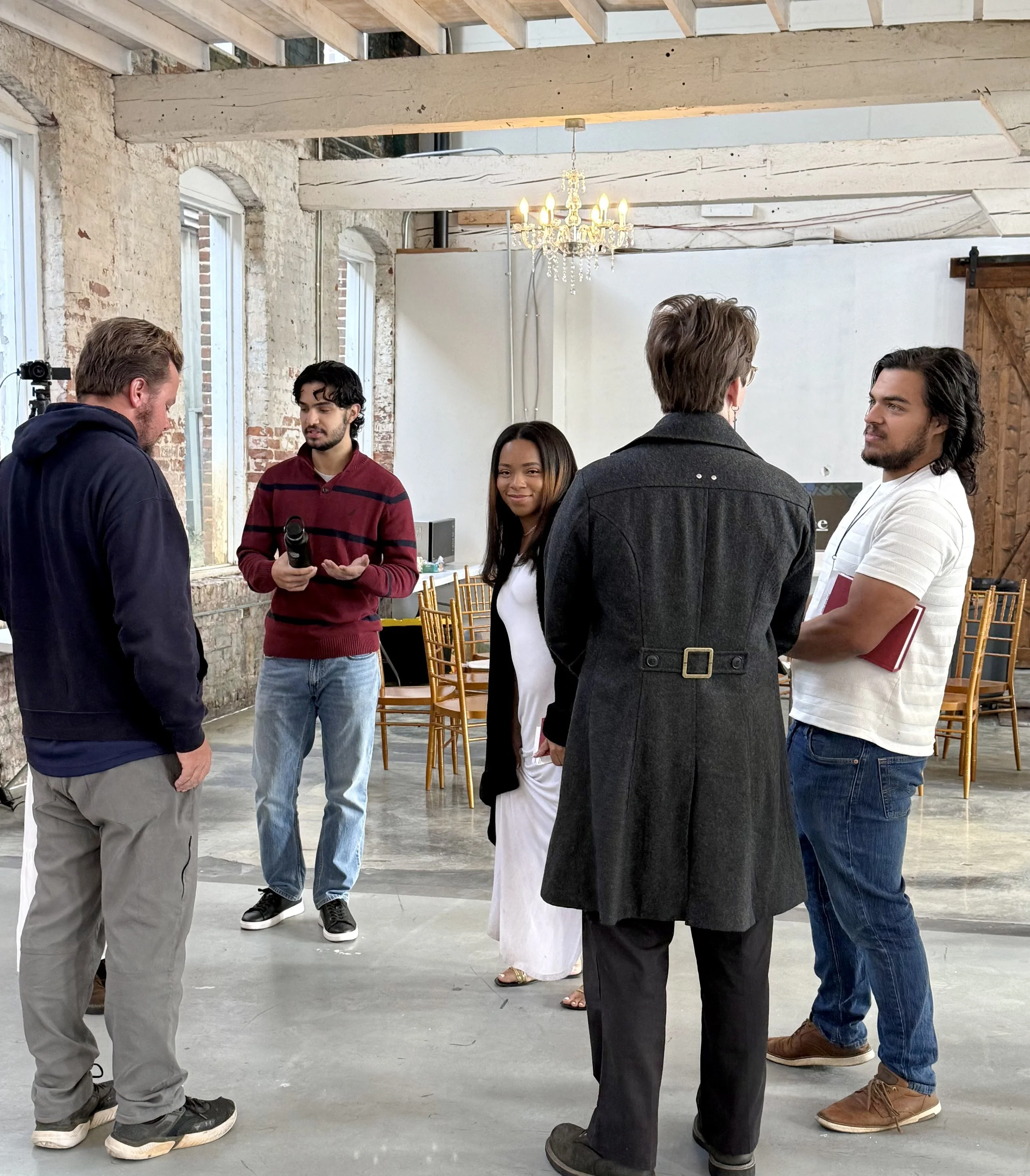 Group of five people having a discussion in a bright industrial-style room with brick walls and large windows, with chairs and a chandelier overhead.
