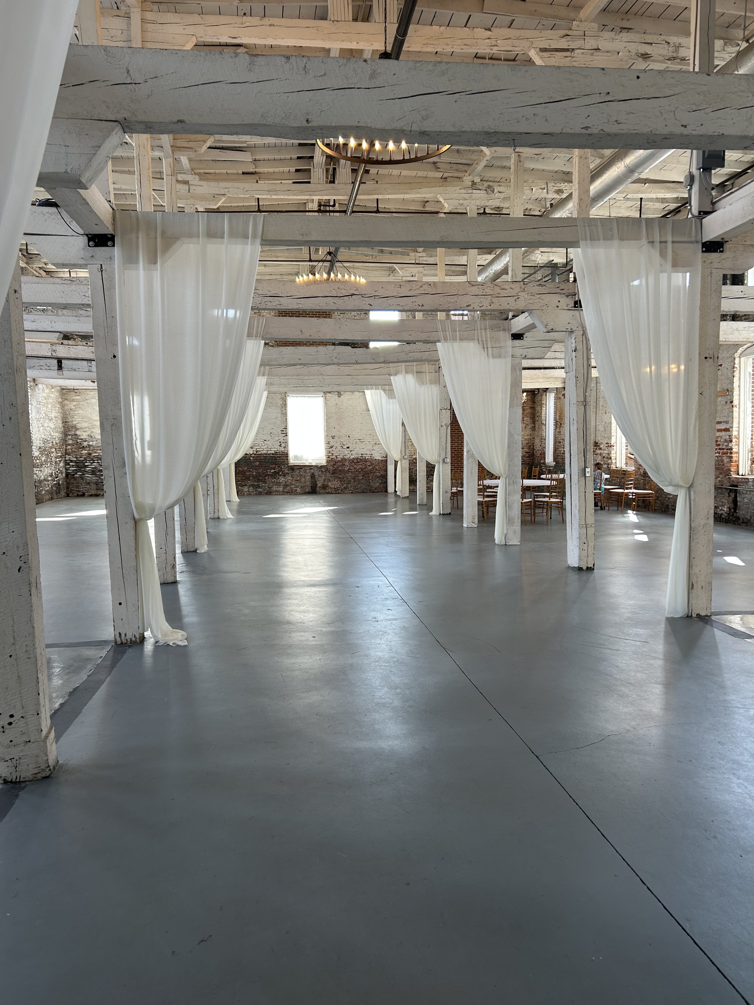 Empty event space with white curtains hanging from wooden beams, concrete floors, brick walls, and large windows.