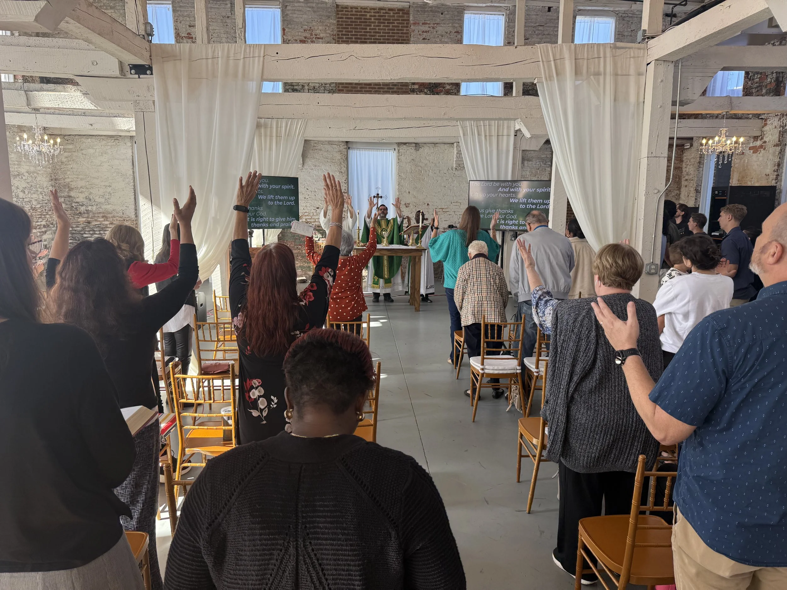 People gathered in a church or worship space, with some raising their hands in prayer or worship, facing a choir and altar at the front with a priest or pastor.