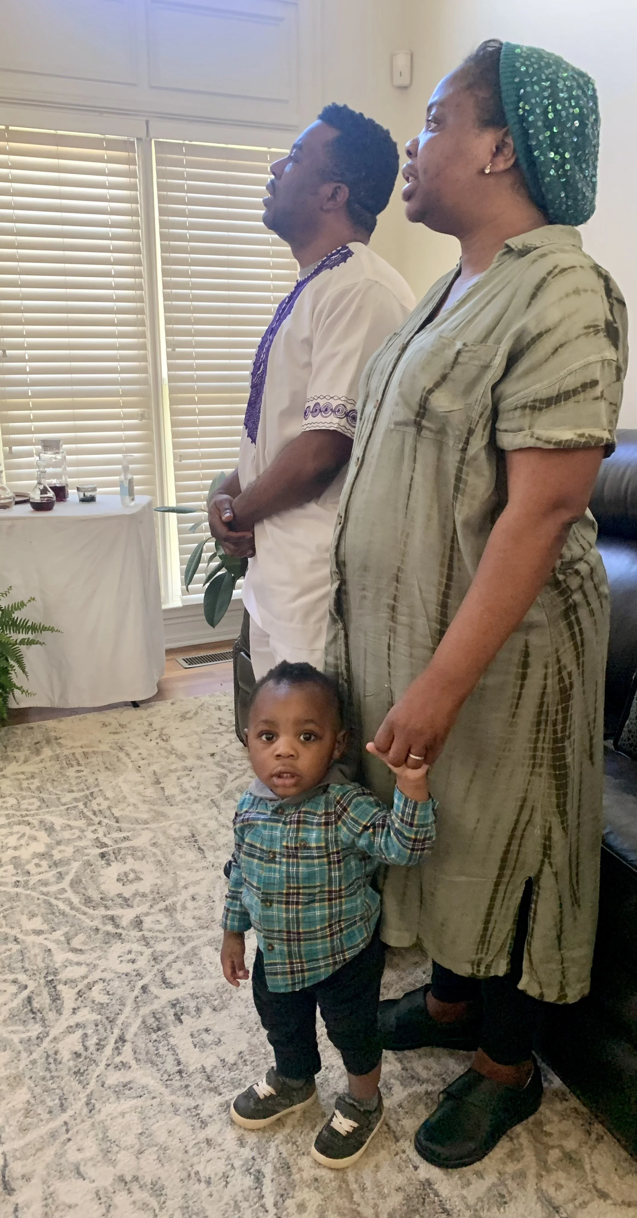 A woman holds a young boy's hand while standing in a living room with patterned carpet, onlookers are praying or singing in the background.