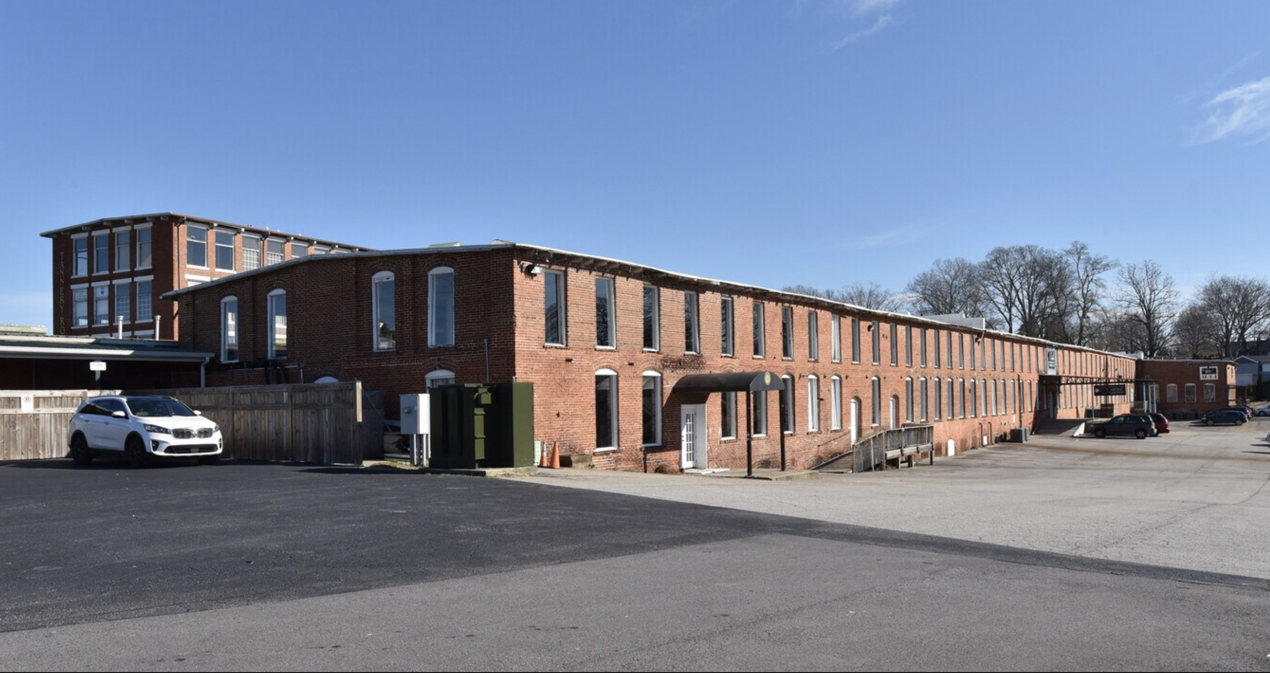A long red brick building with multiple windows, parking lot in the foreground, cars parked along the side, and a clear blue sky above.