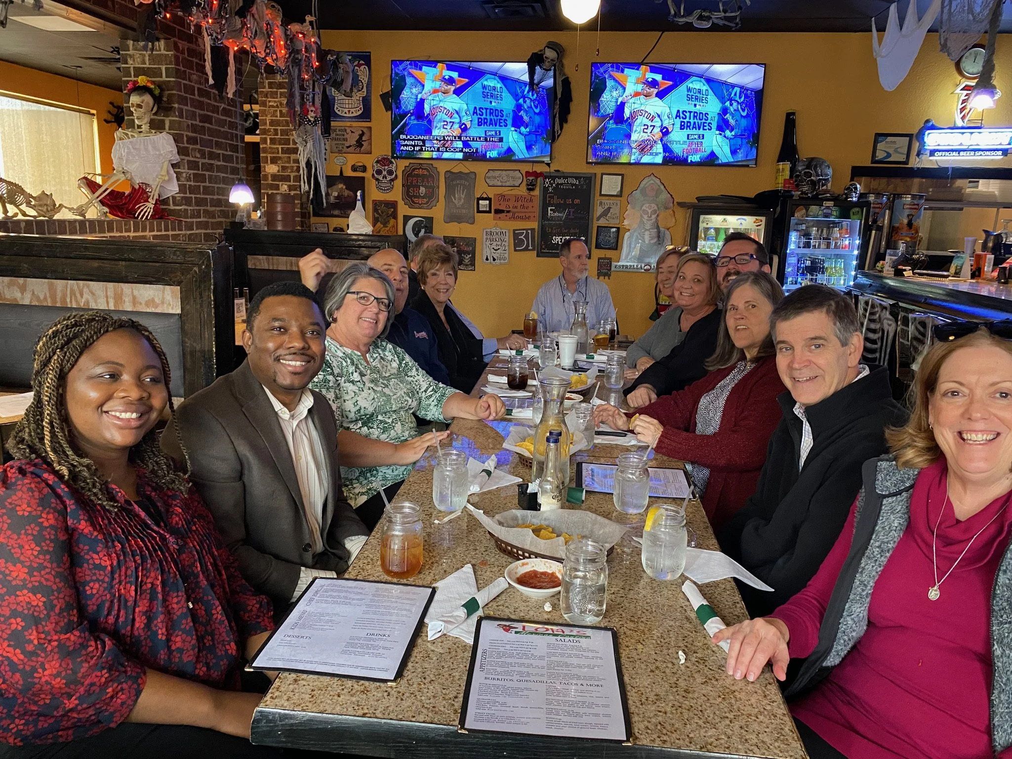 A group of people sitting around a long table inside a restaurant, smiling for a photo. The restaurant has Halloween decorations on the walls, with TV screens showing sports in the background.