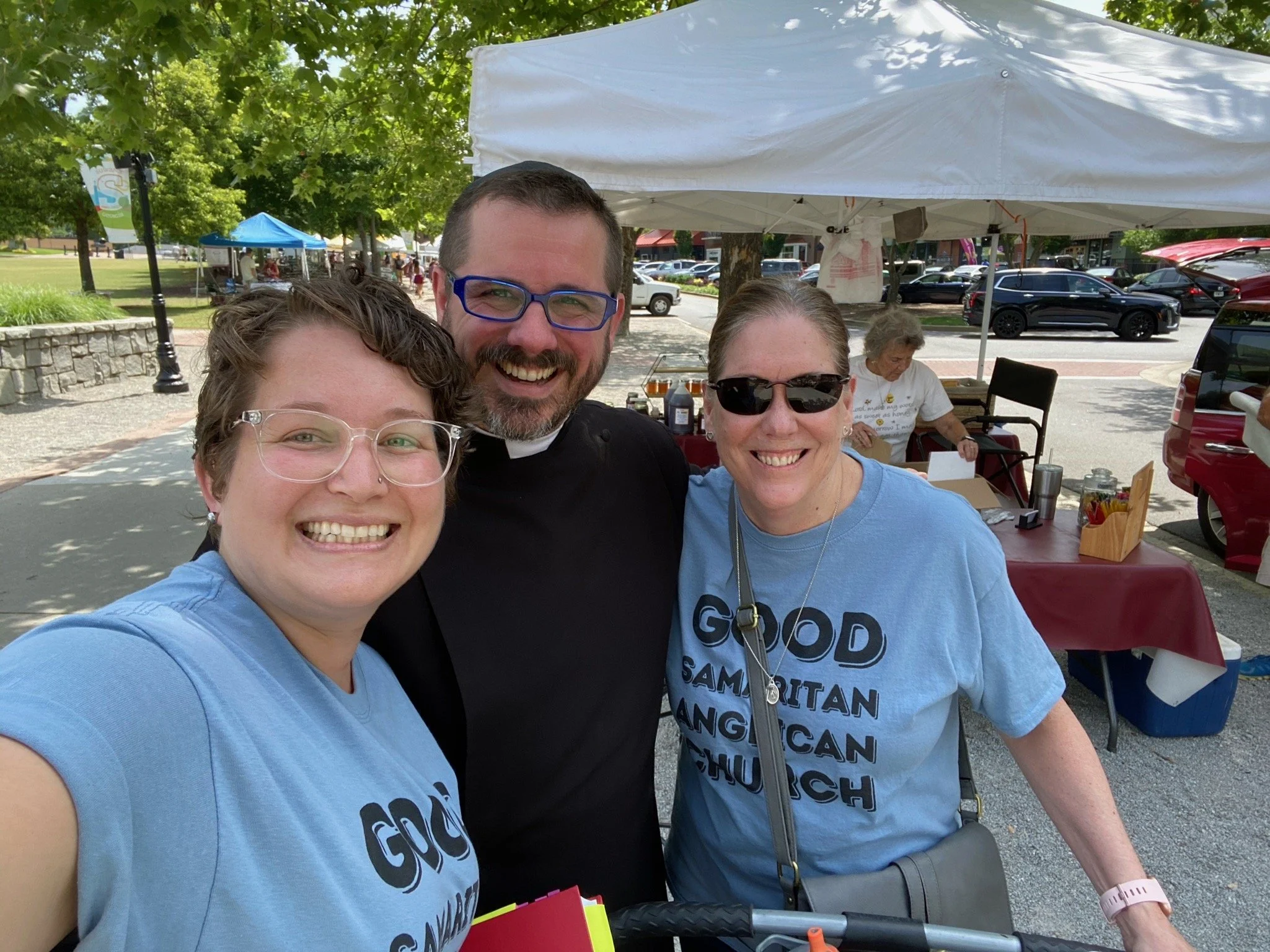 Three smiling people taking a selfie outdoors at a daytime event with tents and parked cars in the background.