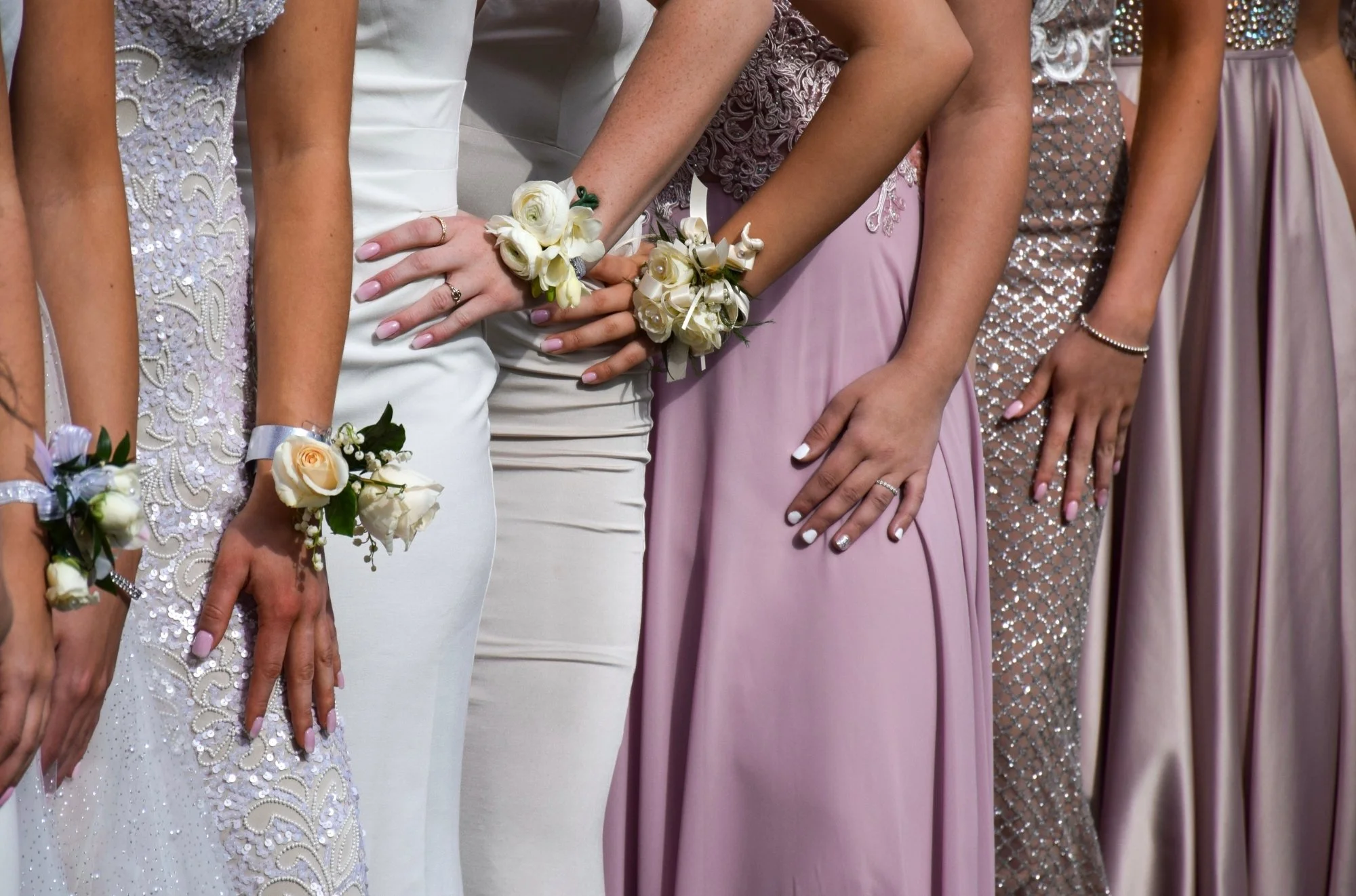 Close-up of a line of women dressed in elegant gowns with corsages on their wrists, standing during a formal event or wedding.
