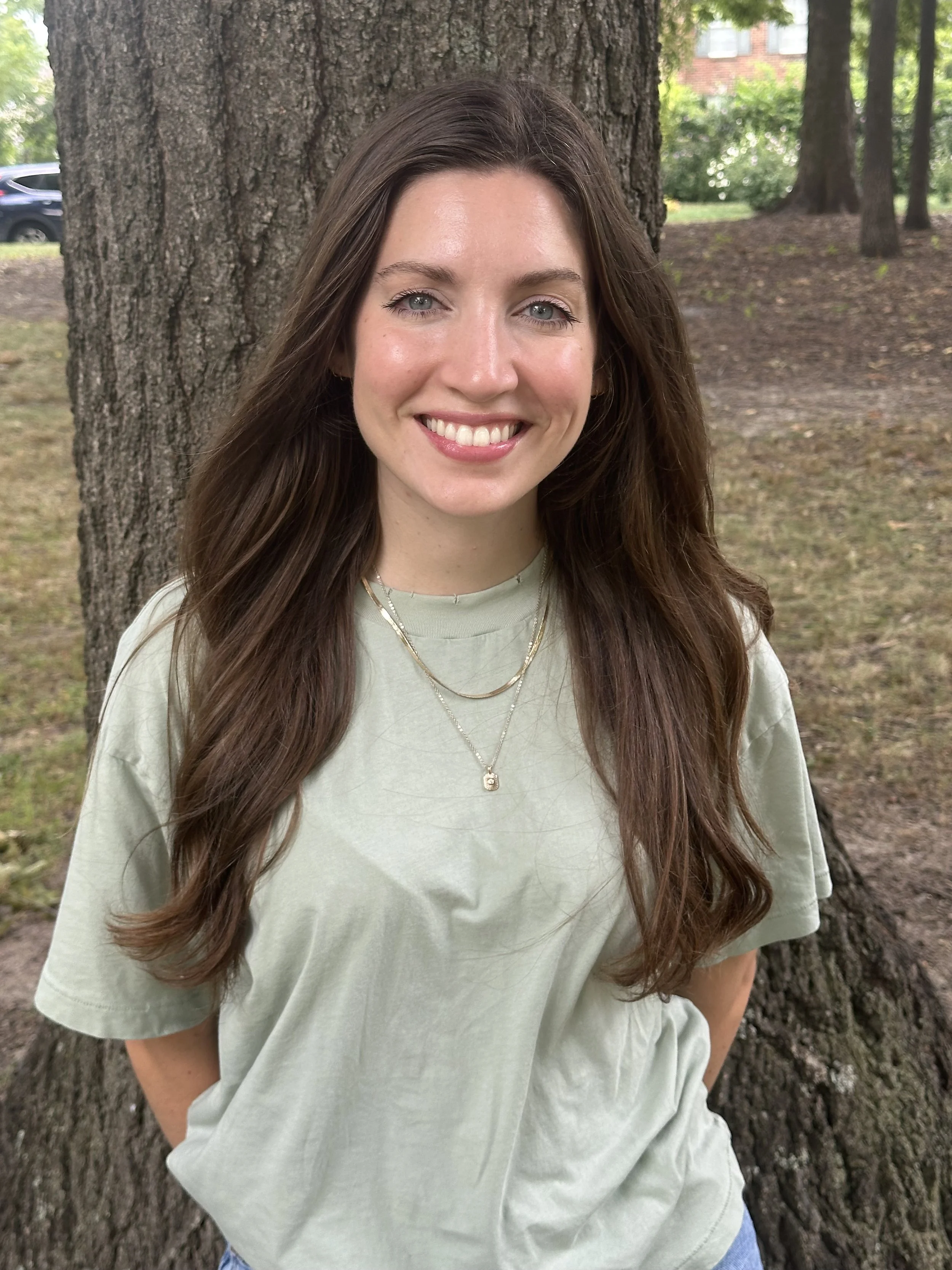 A young woman with long brown hair, smiling, standing outdoors against a large tree in a park, wearing a light green t-shirt and layered necklaces.