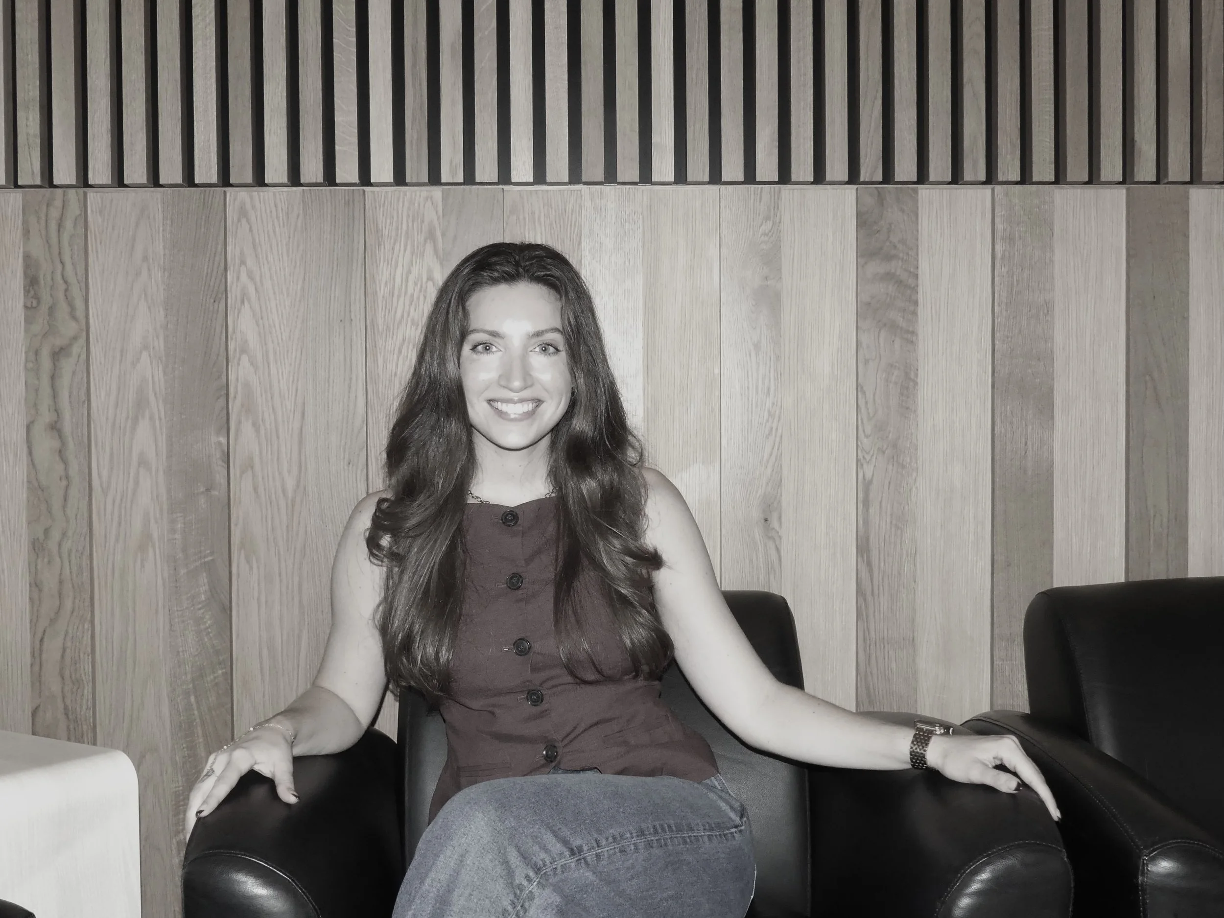 A woman with long, wavy hair sitting at a table in front of a wooden-paneled wall, smiling at the camera.