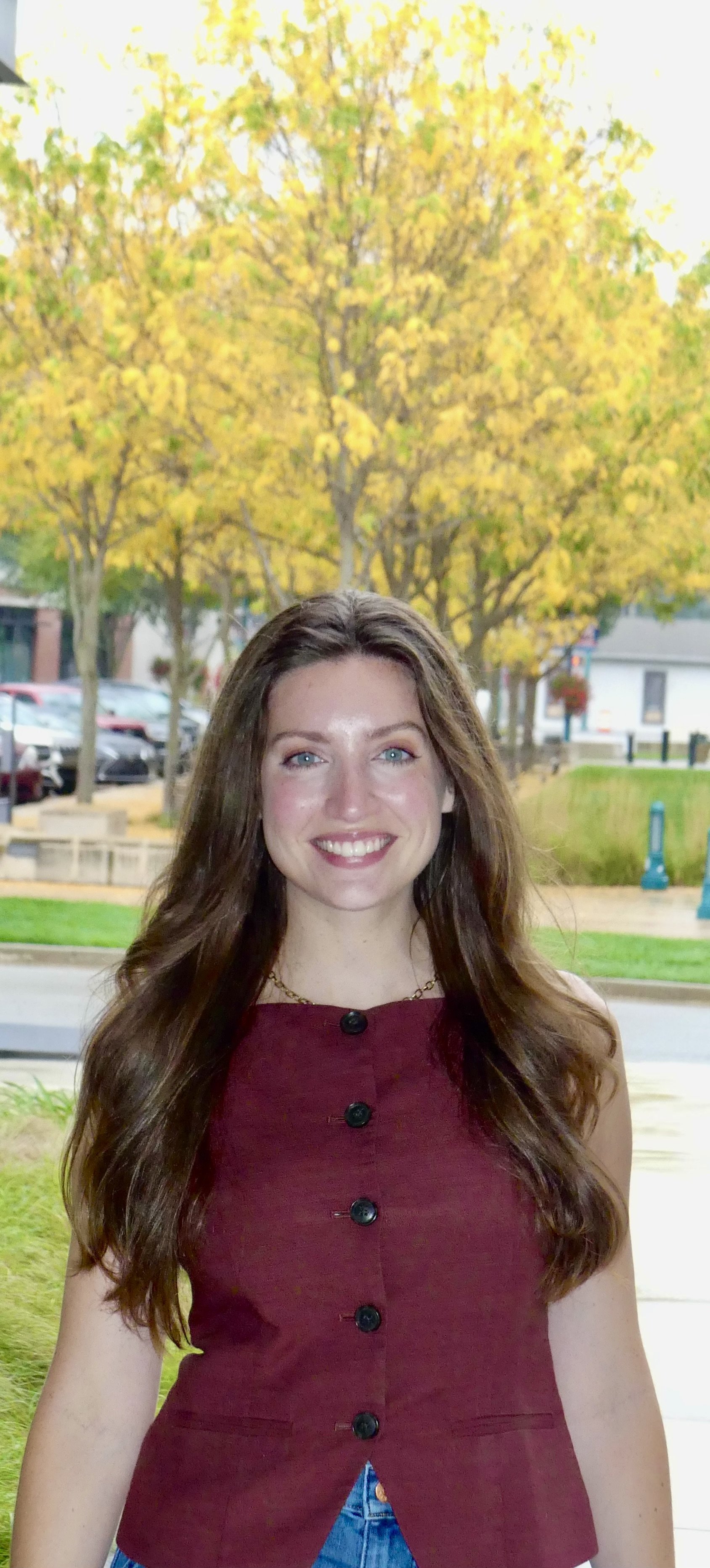 A young woman with long, wavy brown hair and blue eyes smiling outdoors in front of trees with yellow and green leaves, dressed in a sleeveless burgundy top with black buttons and blue jeans.