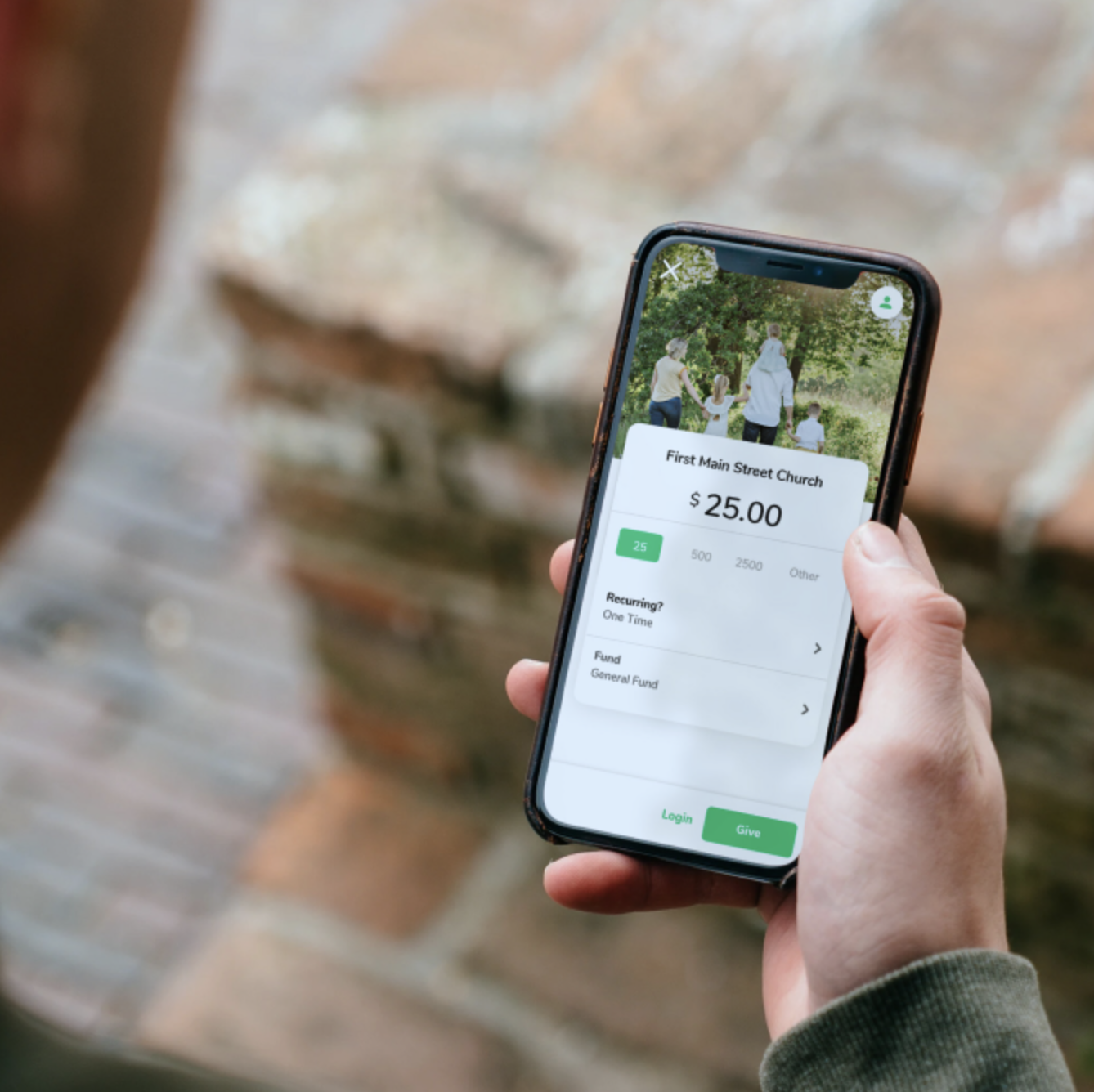 Person holding a smartphone displaying a donation screen for First Main Street Church with a $25 donation option, an image of people walking in a park, and donation details such as recurring and fund options.