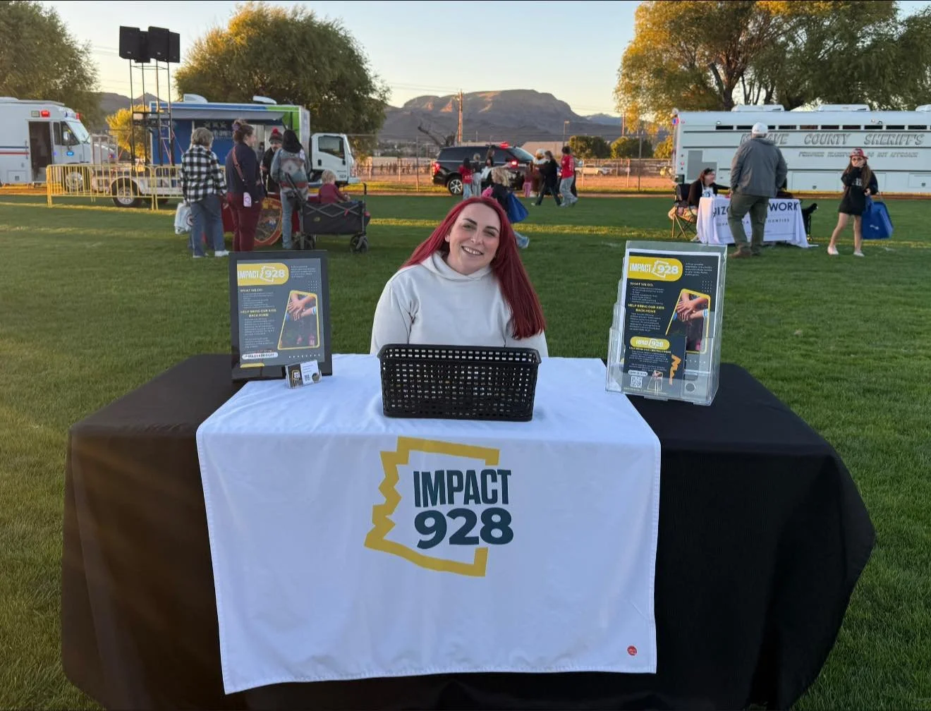 A woman with red hair smiling and sitting at a table with a white banner that reads IMPACT 928, set up outdoors on a grassy field during a community event. The table has brochures and informational materials, and there are other booths and people in the background.