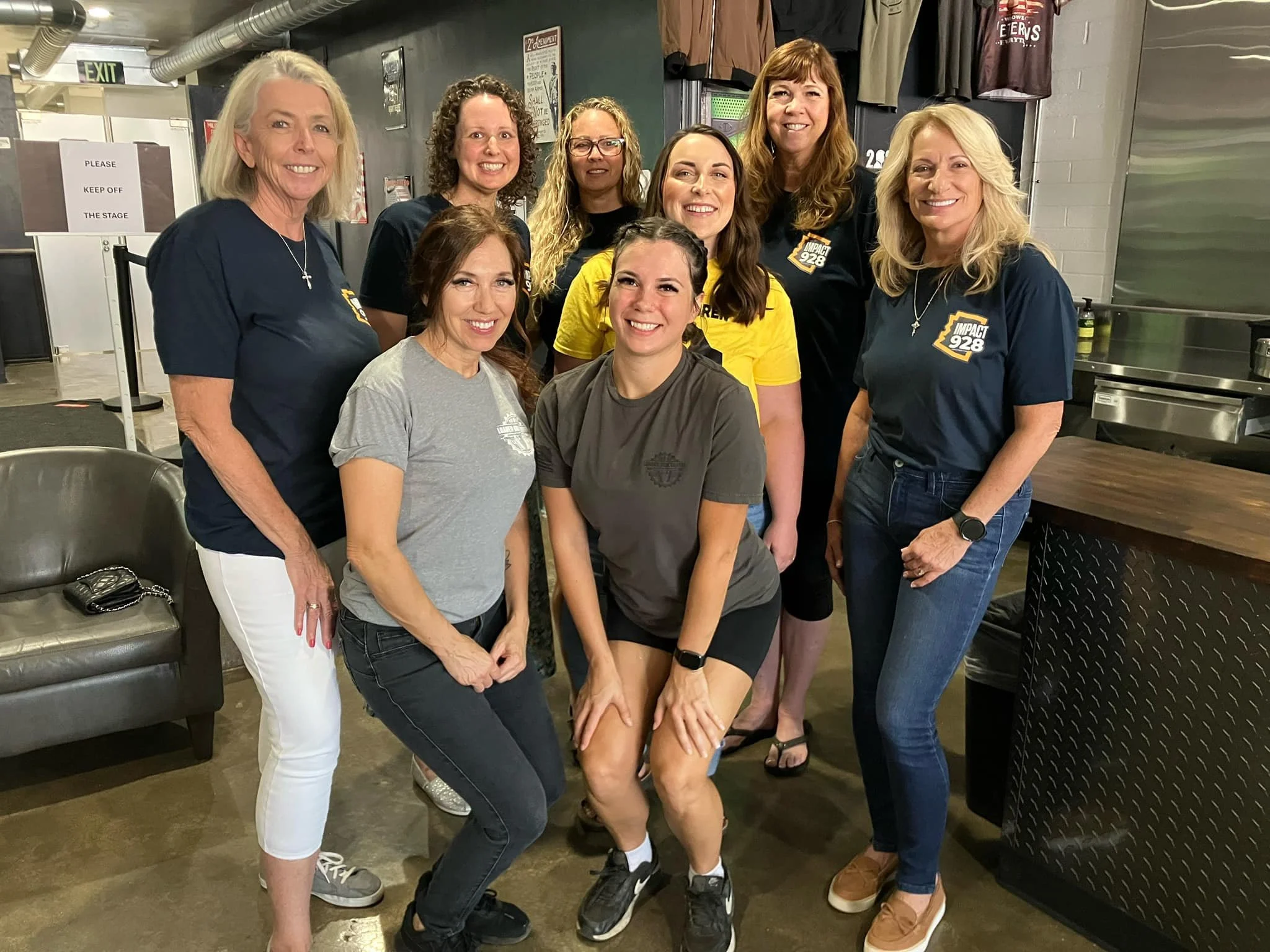 Group of nine women posing together indoors, some wearing T-shirts with 'Impact 928' logo, smiling at the camera.