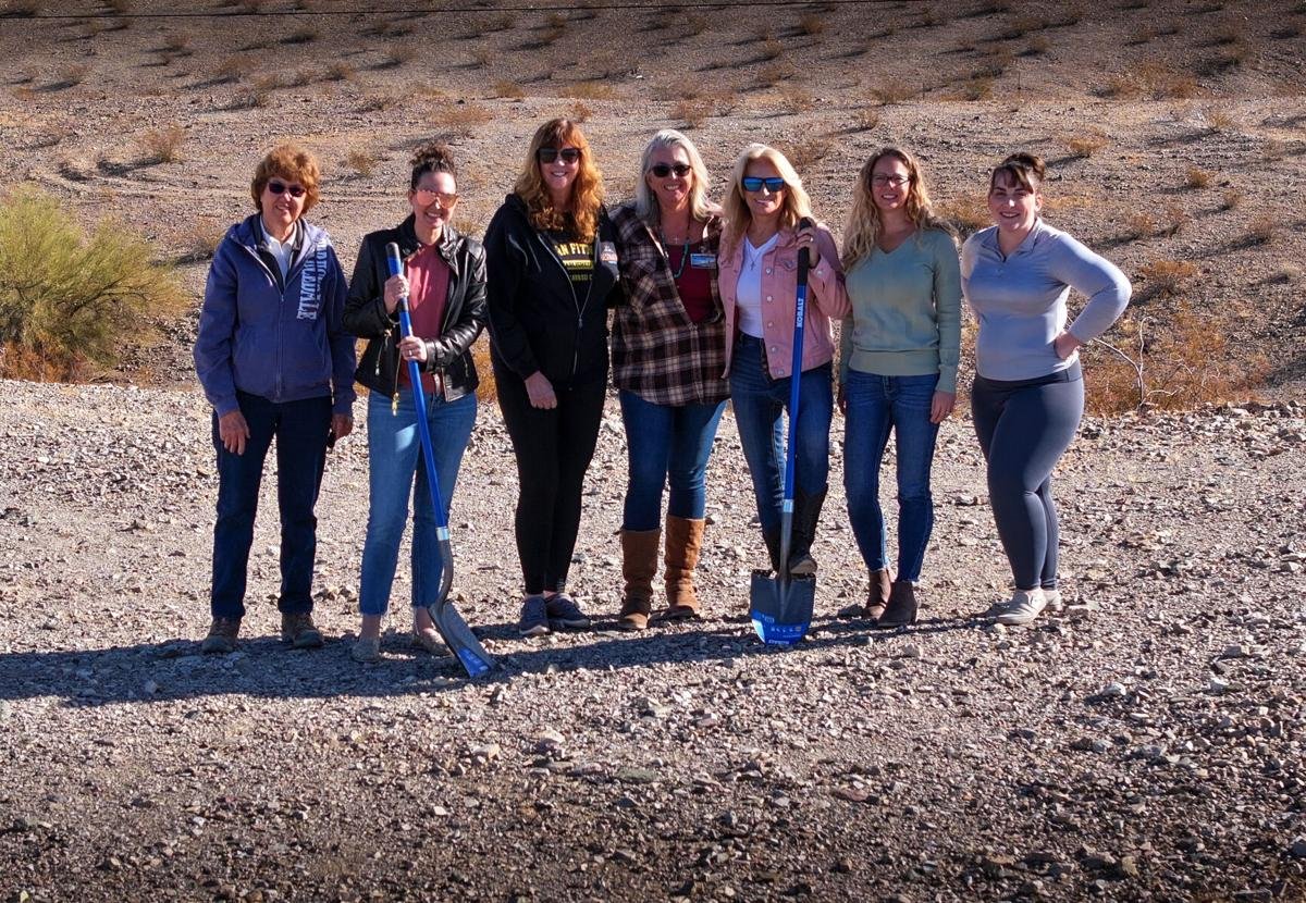 Six women standing together on a gravel path in a desert landscape, smiling at the camera, some holding shovels, dressed casually for outdoor activity.