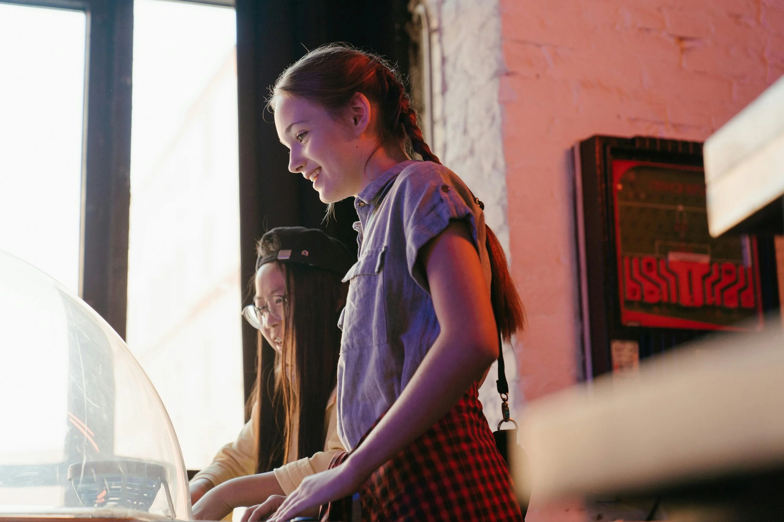 Two young women are playing arcade games together, smiling and focused. The girl in the foreground has long hair in a braid, wearing a short-sleeved shirt and a red checkered skirt; the girl behind her has long straight hair, glasses, and a cap. They are indoors, with neon lighting and an arcade game visible.