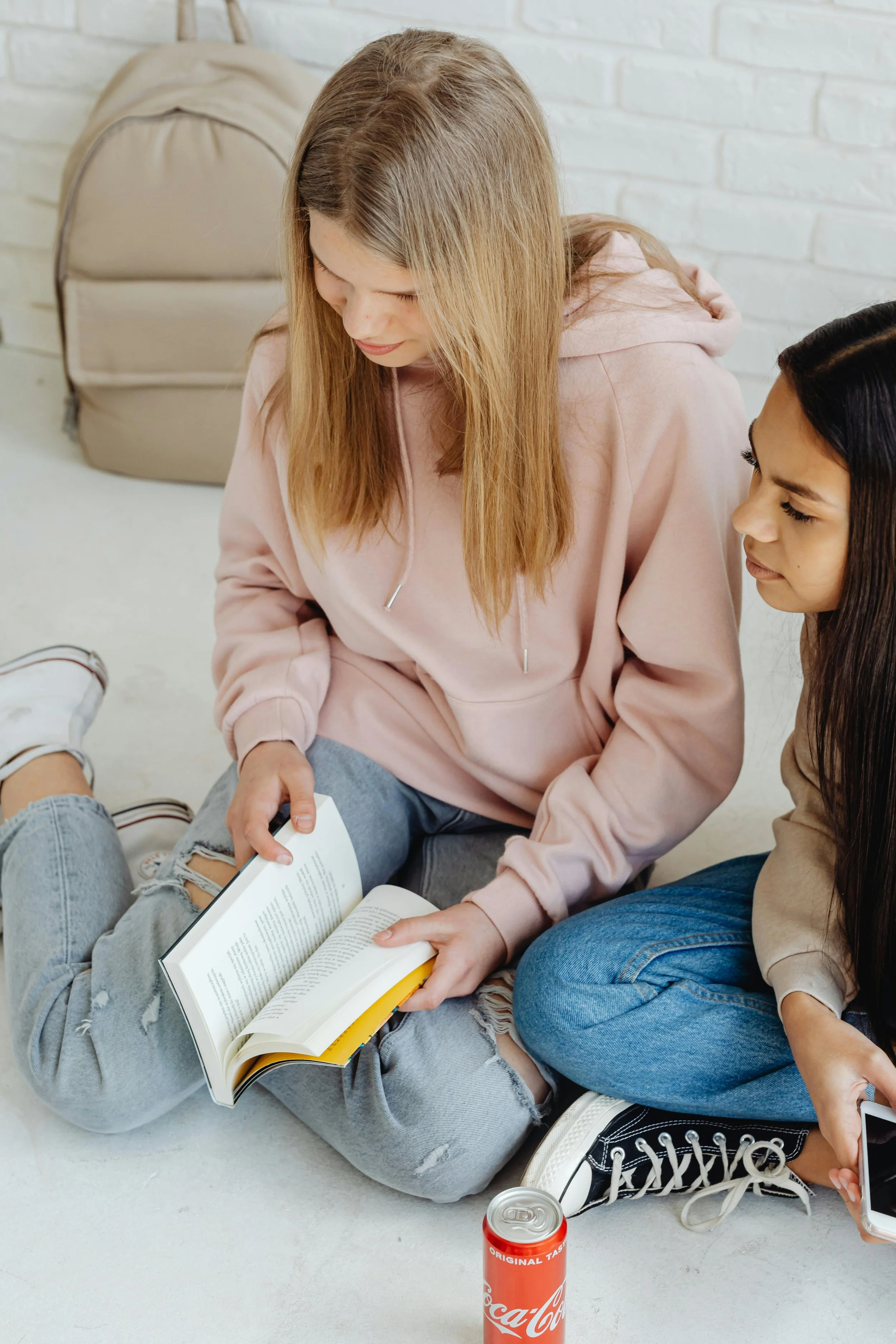 Two teenage girls sitting on the floor, one reading a book and the other holding a phone, with a backpack and a soda can nearby.