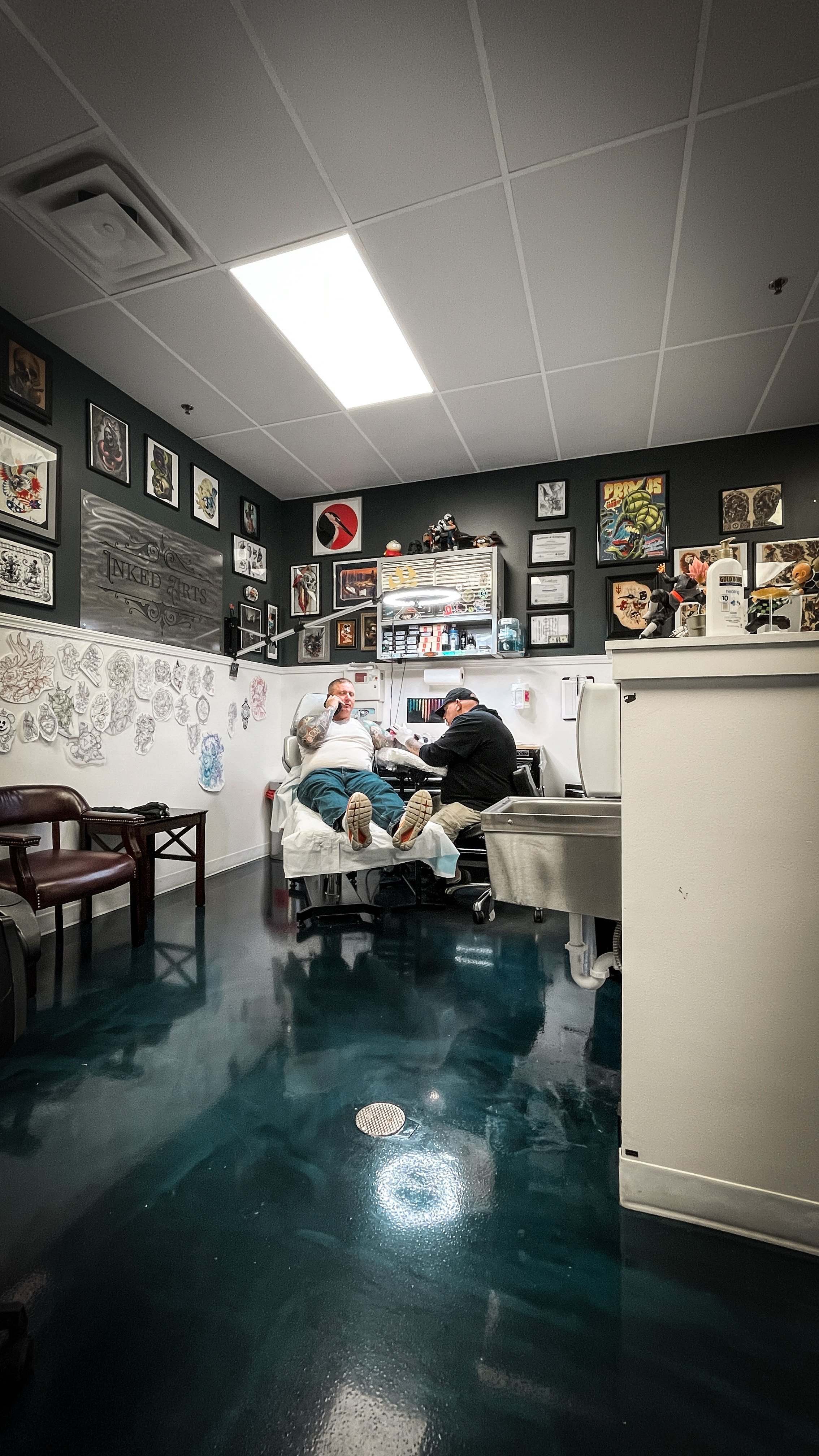 Tattoo artist working on a tattoo of a man lying on a chair in a tattoo studio decorated with framed artwork and tattoos on the wall.