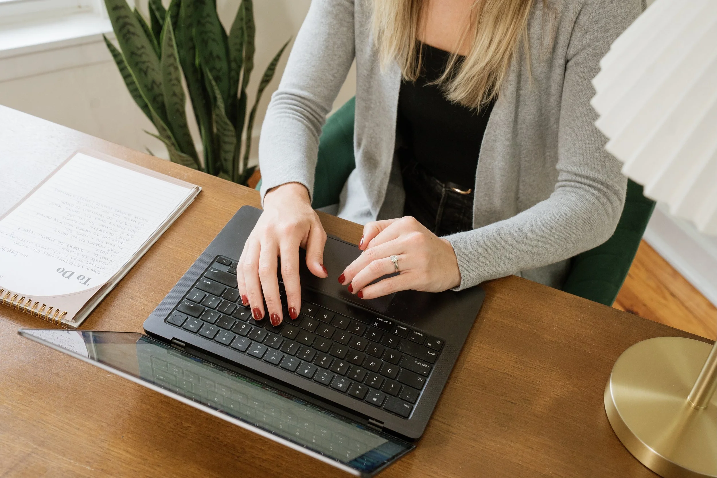 A woman working on a laptop at a desk with a notebook to her left and a gold lamp to her right. There is a green plant in the background.