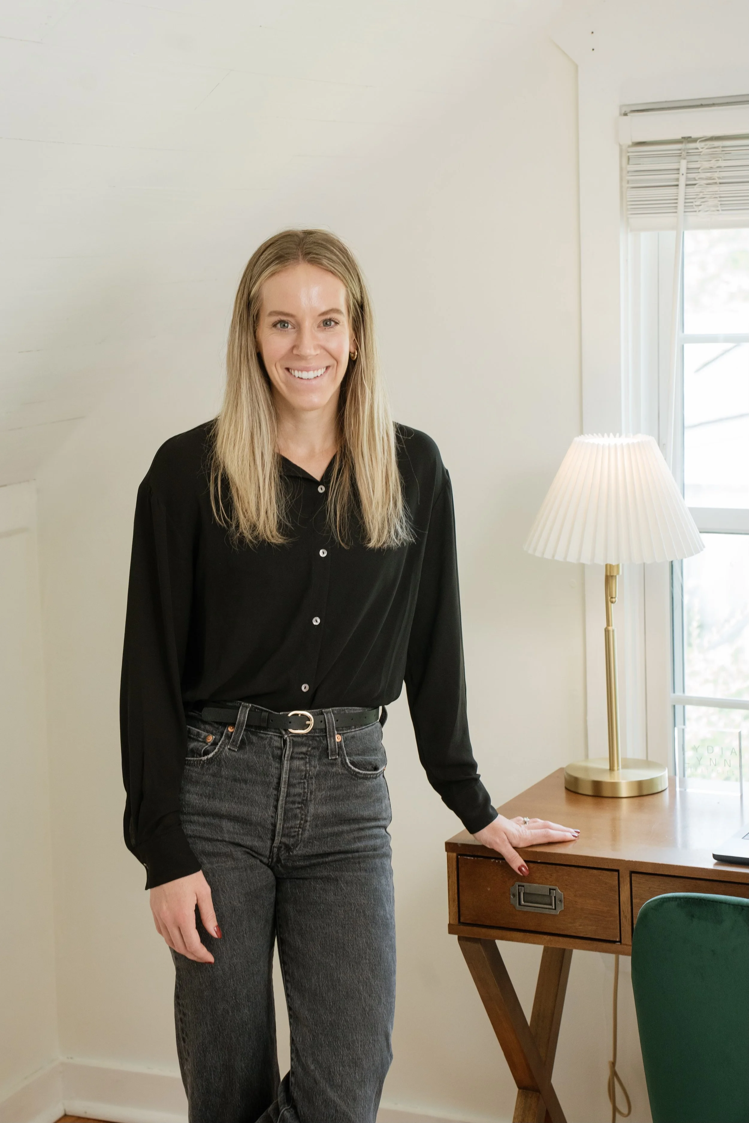 A woman with long blonde hair wearing a black blouse and gray jeans, standing next to a wooden desk with a cream-colored lamp, in a room with a window and white walls.