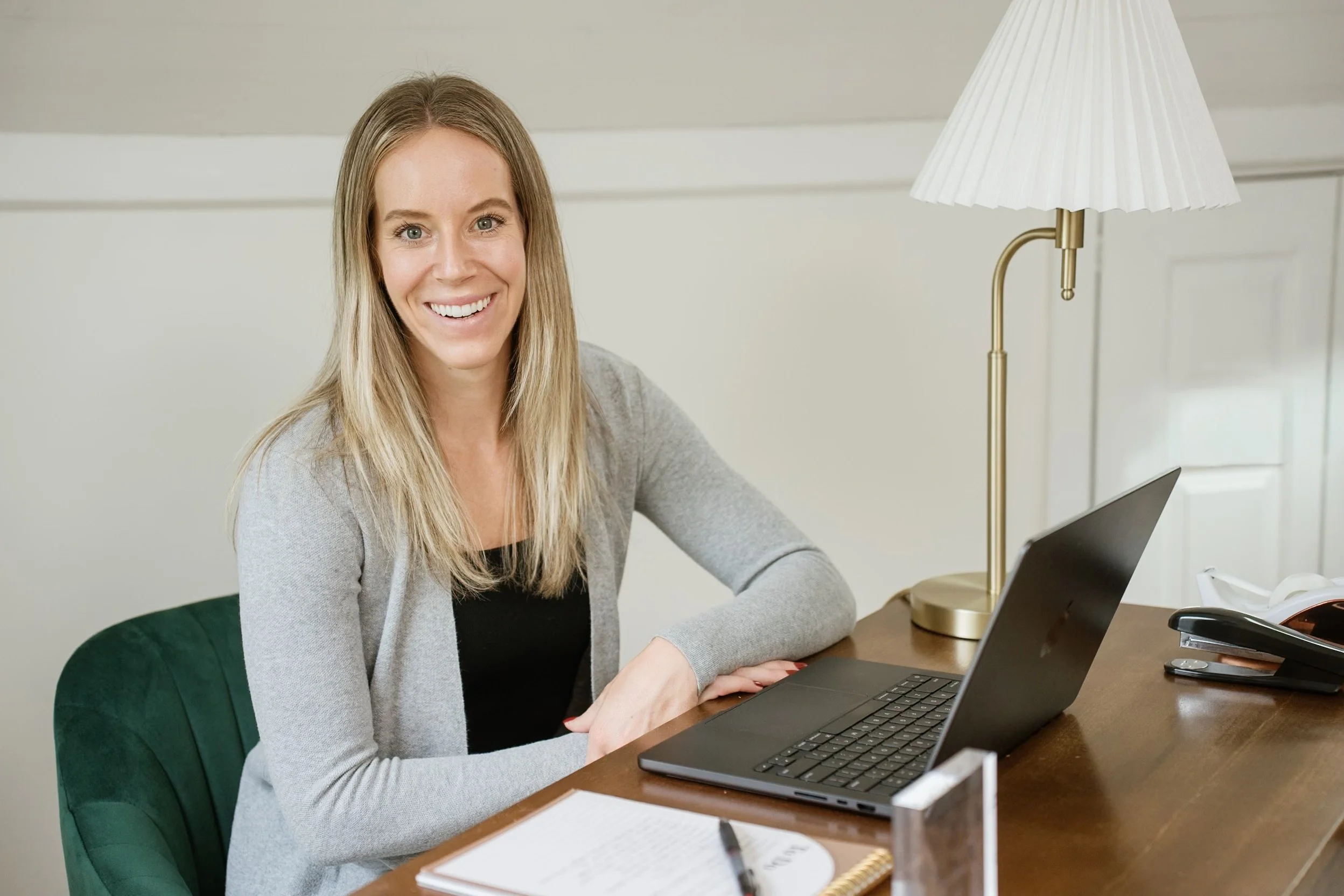 A smiling woman with blonde hair sitting at a desk with a laptop, notepad, pen, and a desk lamp.