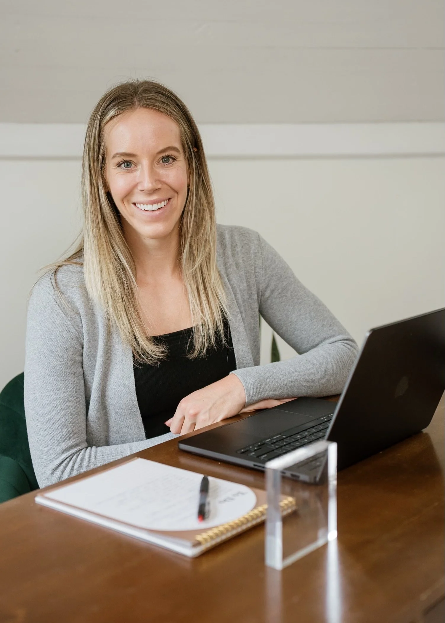 A woman sitting at a wooden desk with a black laptop, smiling, in a professional office setting, with a notebook and pen in front of her.