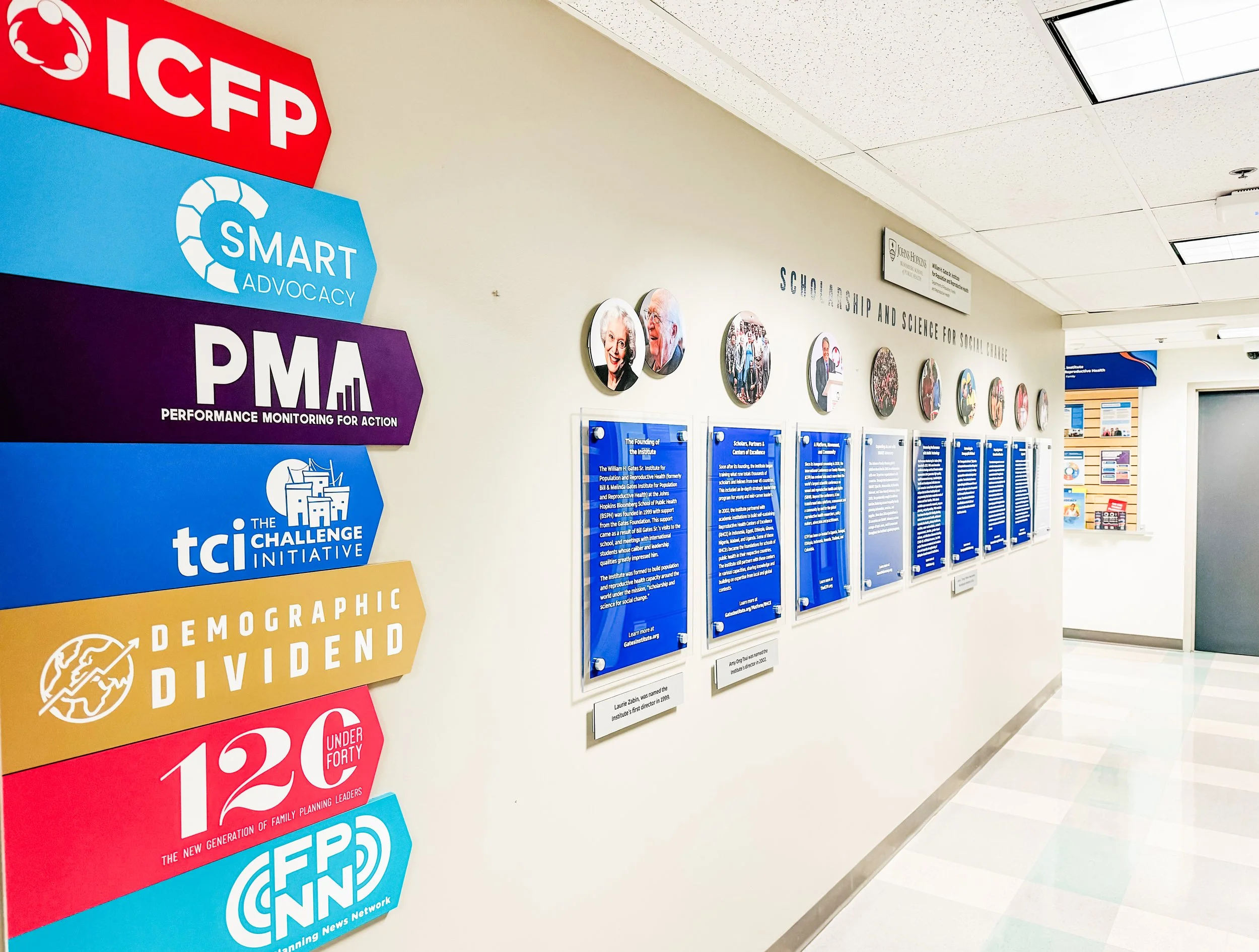 Hospital hallway with colorful signs on the wall, including logos and phrases such as ICFP, SMART Advocacy, PMA, TCI Initiative, and Demographic Division, along with framed photographs and informational posters.