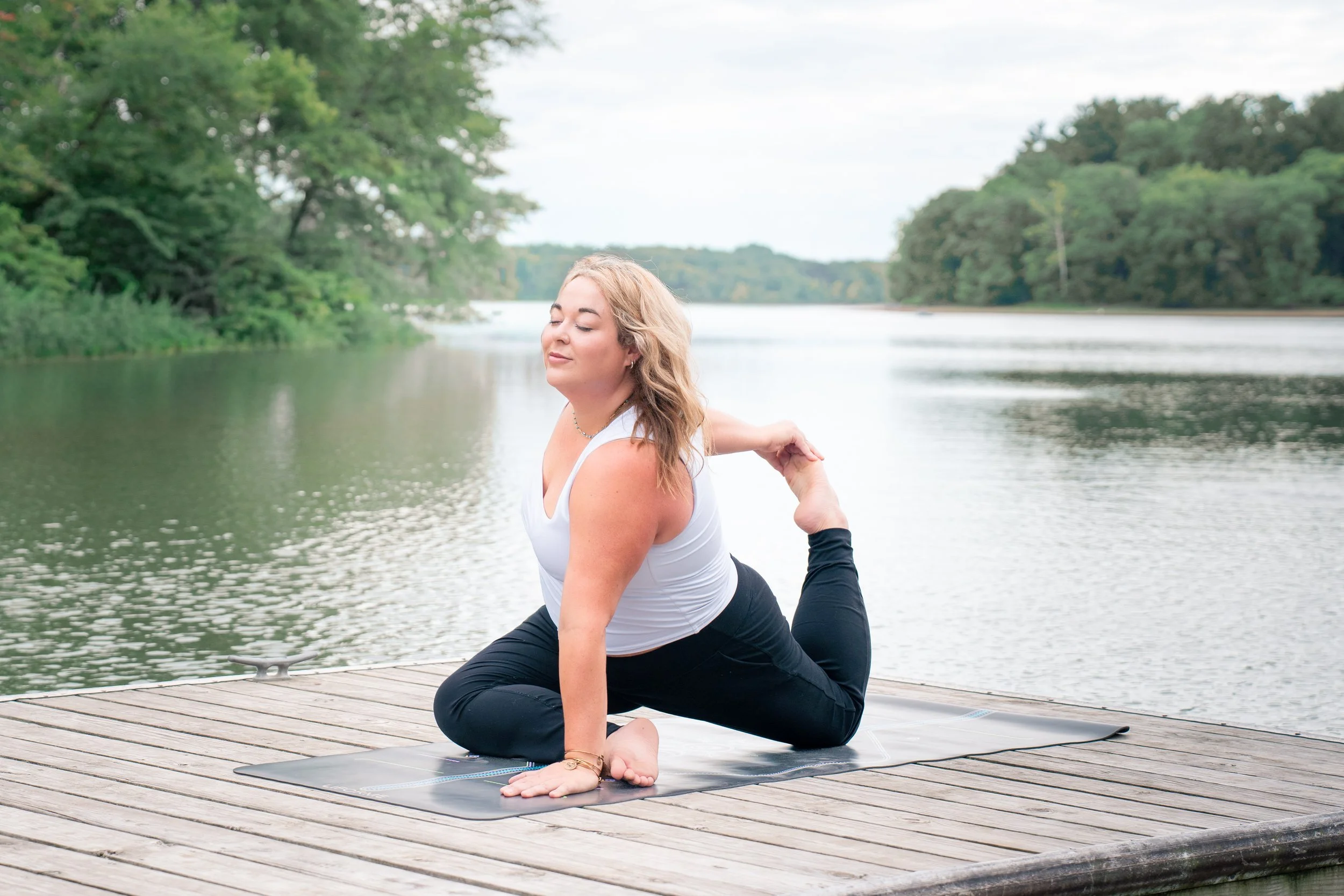 A woman practicing yoga on a dock beside a lake, performing a backbend stretch with one hand on her ankle and the other on the ground, surrounded by lush green trees.