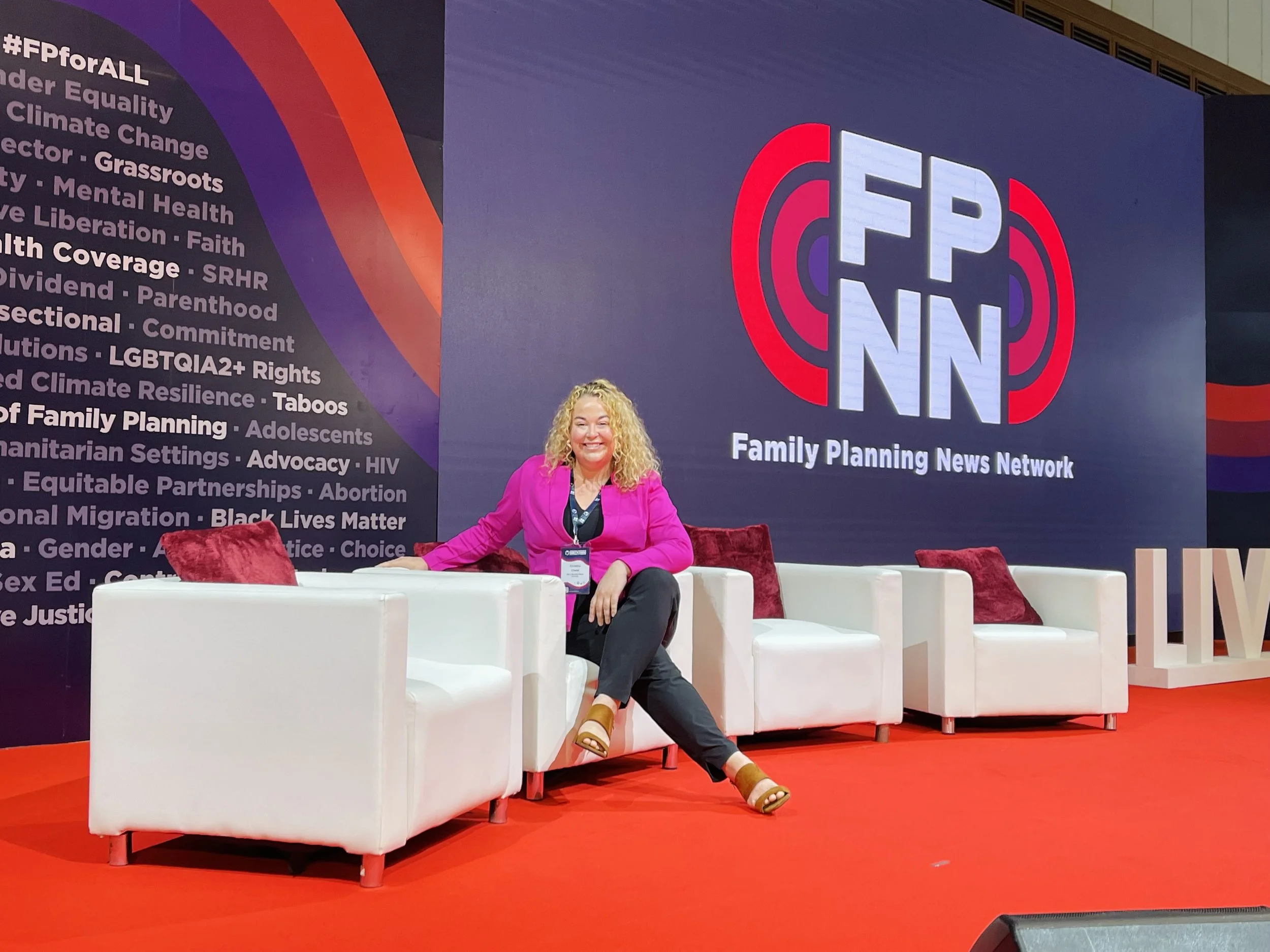 A woman with blonde curly hair, wearing a pink blazer, black pants, and brown sandals, smiling while sitting on a white chair at a conference or event. Behind her is a large sign with the logo of Family Planning News Network and text about social issues such as equality, climate change, mental health, and reproductive rights, in front of a blue and red background.
