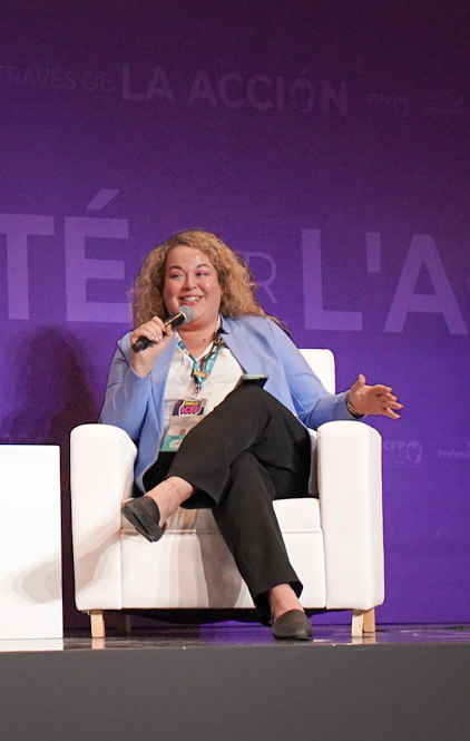 A woman with curly hair, wearing a blue blazer and black pants, sits on a white armchair on a stage, smiling and speaking into a microphone.