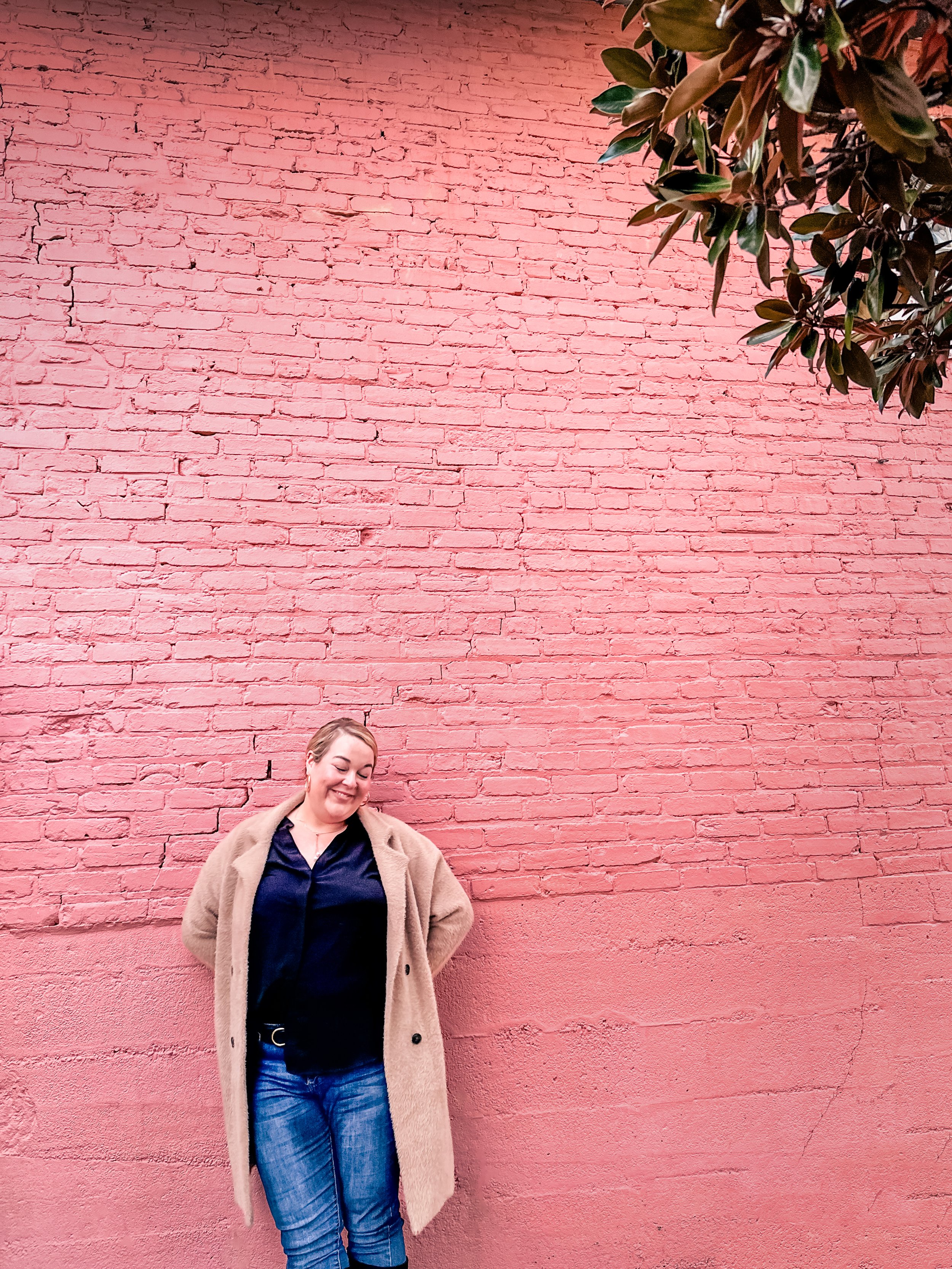 A woman standing in front of a pink brick wall, smiling, wearing a beige coat, dark shirt, and blue jeans, with a large green leafy plant hanging from the top right corner.