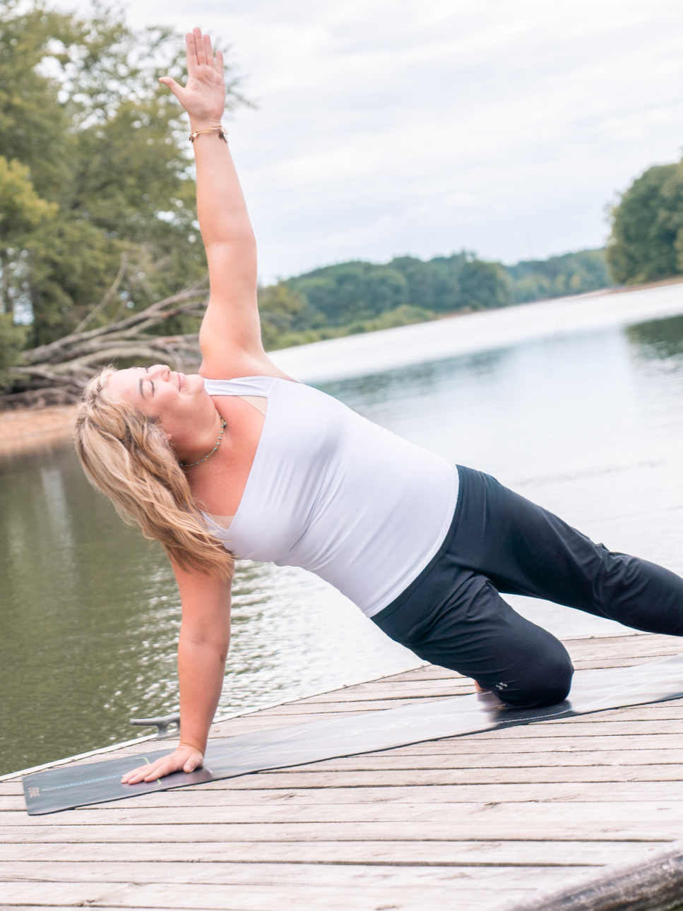 Woman practicing yoga outdoors on a wooden dock by a lake, performing a side plank pose with one arm raised.