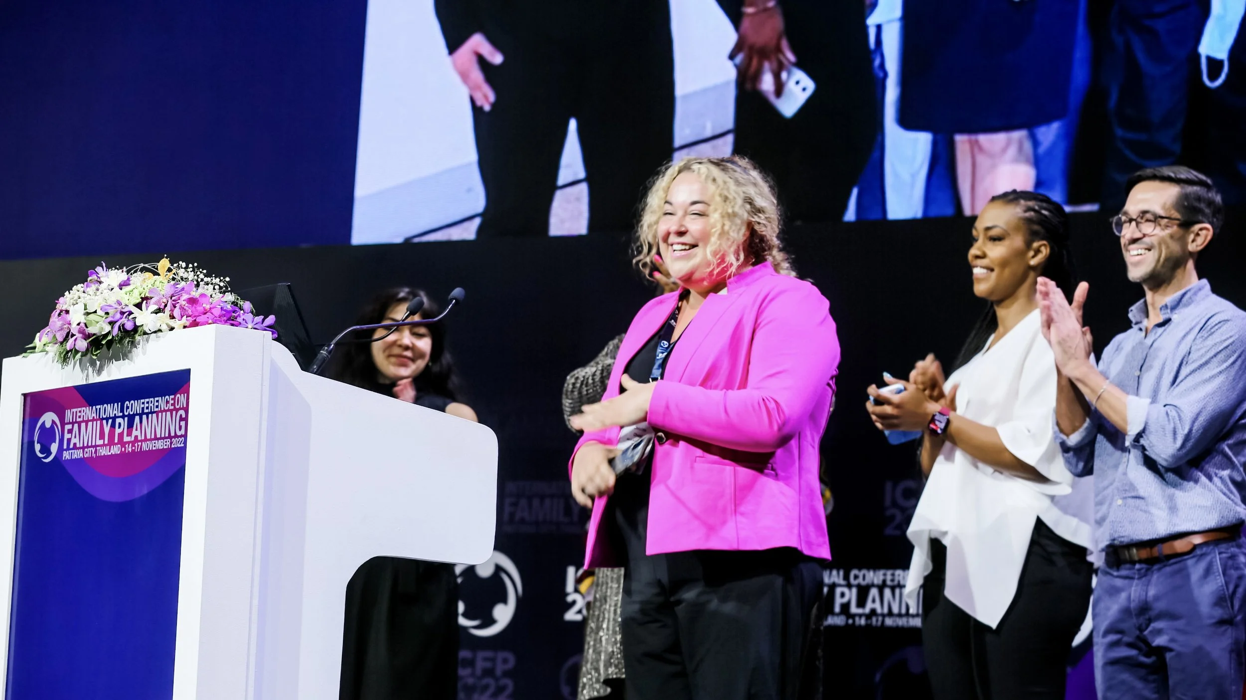 Group of four diverse people on stage at the International Conference on Family Planning, Pattaya, Thailand, November 2022, clapping and smiling, with a podium decorated with flowers and a large screen behind them.