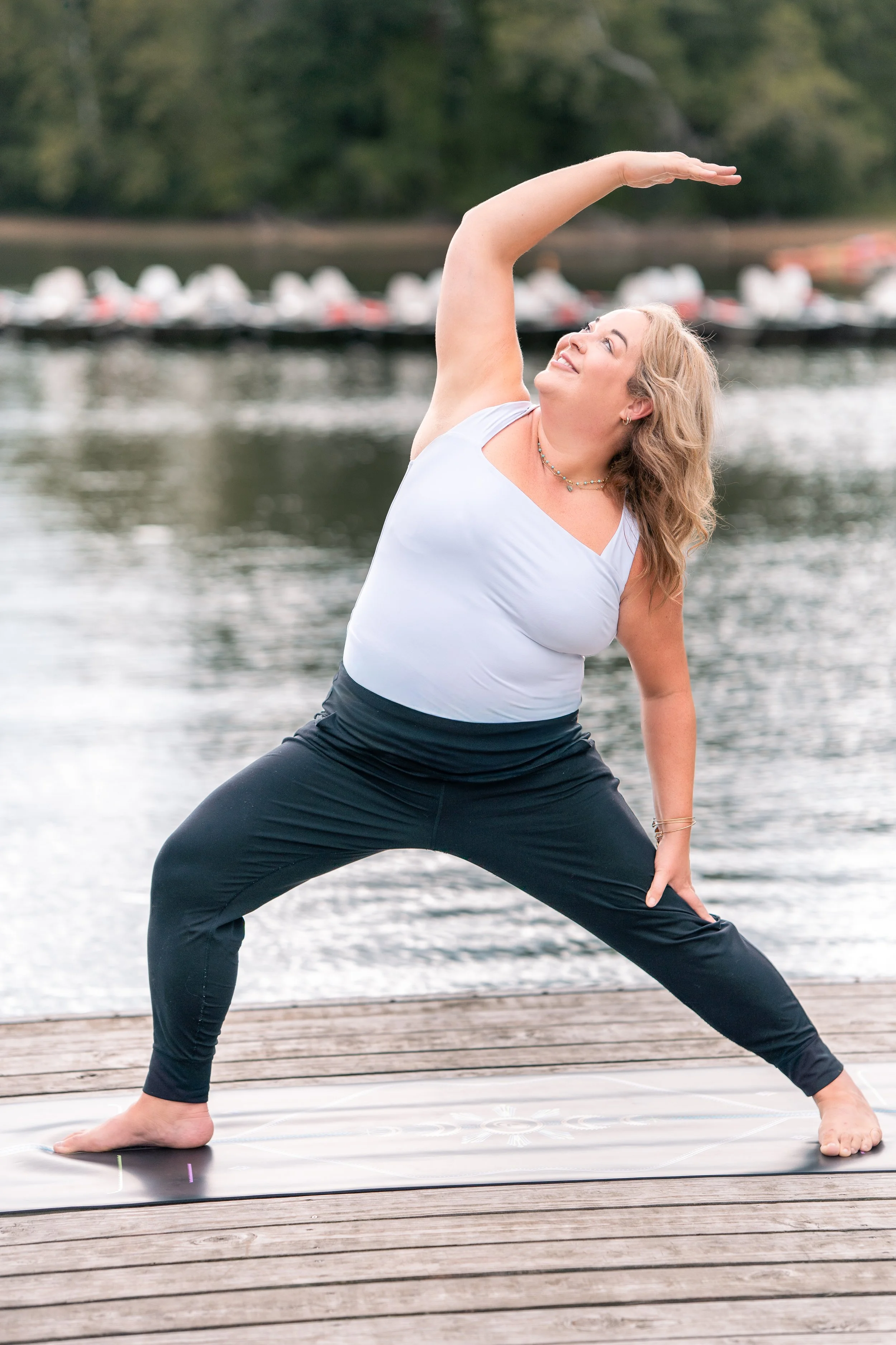 A woman practicing yoga outdoors on a wooden dock near water, performing the Warrior pose with a scenic backdrop of trees and a blurred group of boats or kayaks in the water.