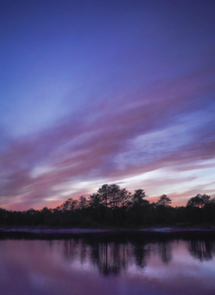 Colorful sunset over a calm lake with silhouetted trees and clouds in the sky.
