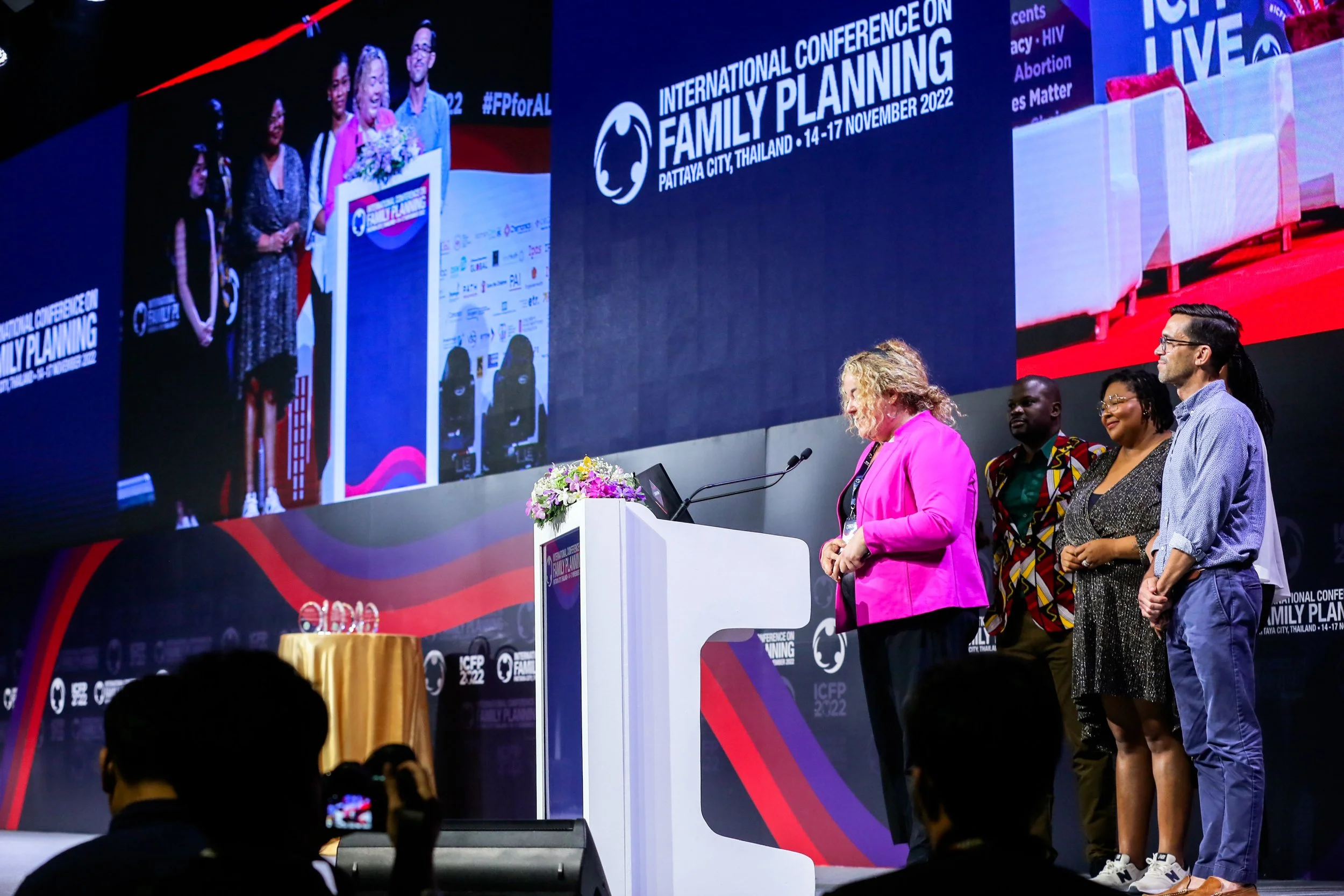 At the International Conference on Family Planning in Pattaya City, Thailand, a panel of diverse speakers stands on stage while a large screen behind displays the event's banner, date, and location. Audience members are visible in the foreground, some recording the event.