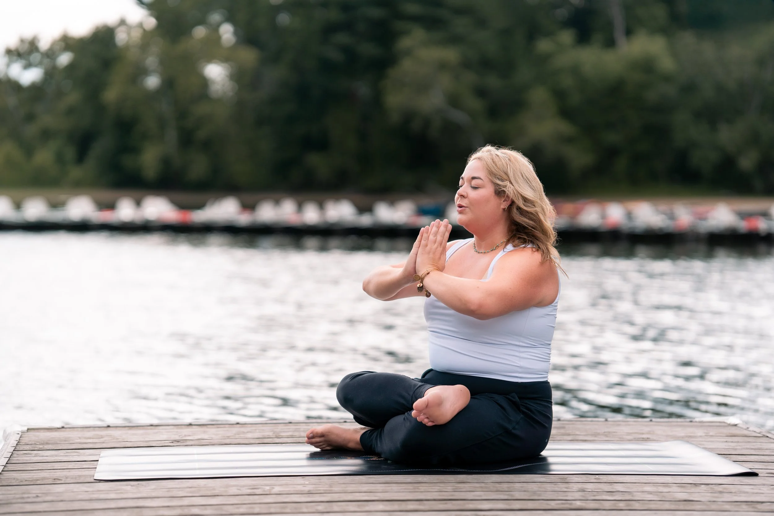 A woman practicing yoga outdoors on a dock by the water, sitting in a cross-legged position with hands in prayer position at her chest, eyes closed.