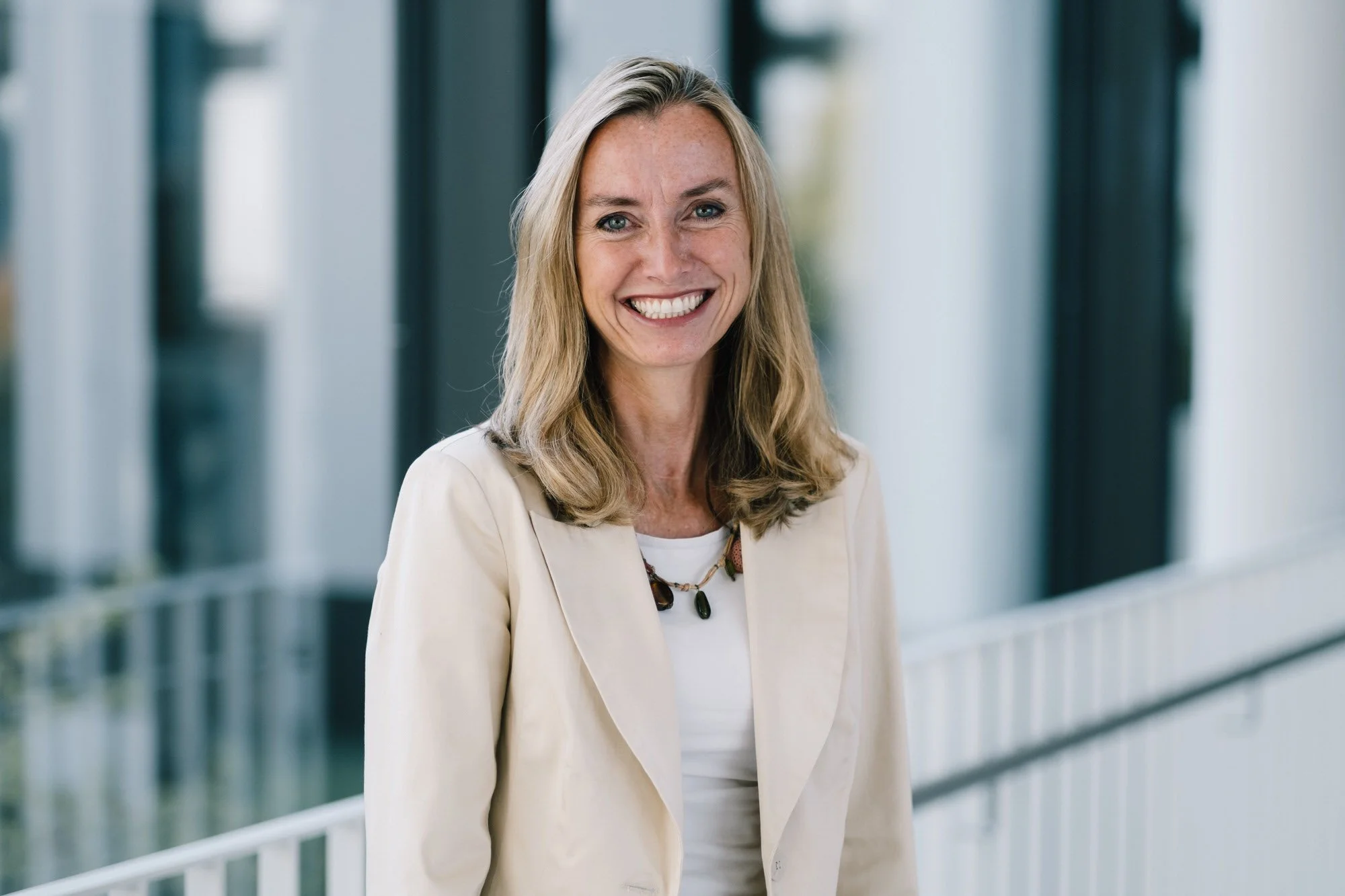 Lächelnde Frau in einem weißen Blazer steht im modernen Gebäude mit großen Fenstern im Hintergrund.