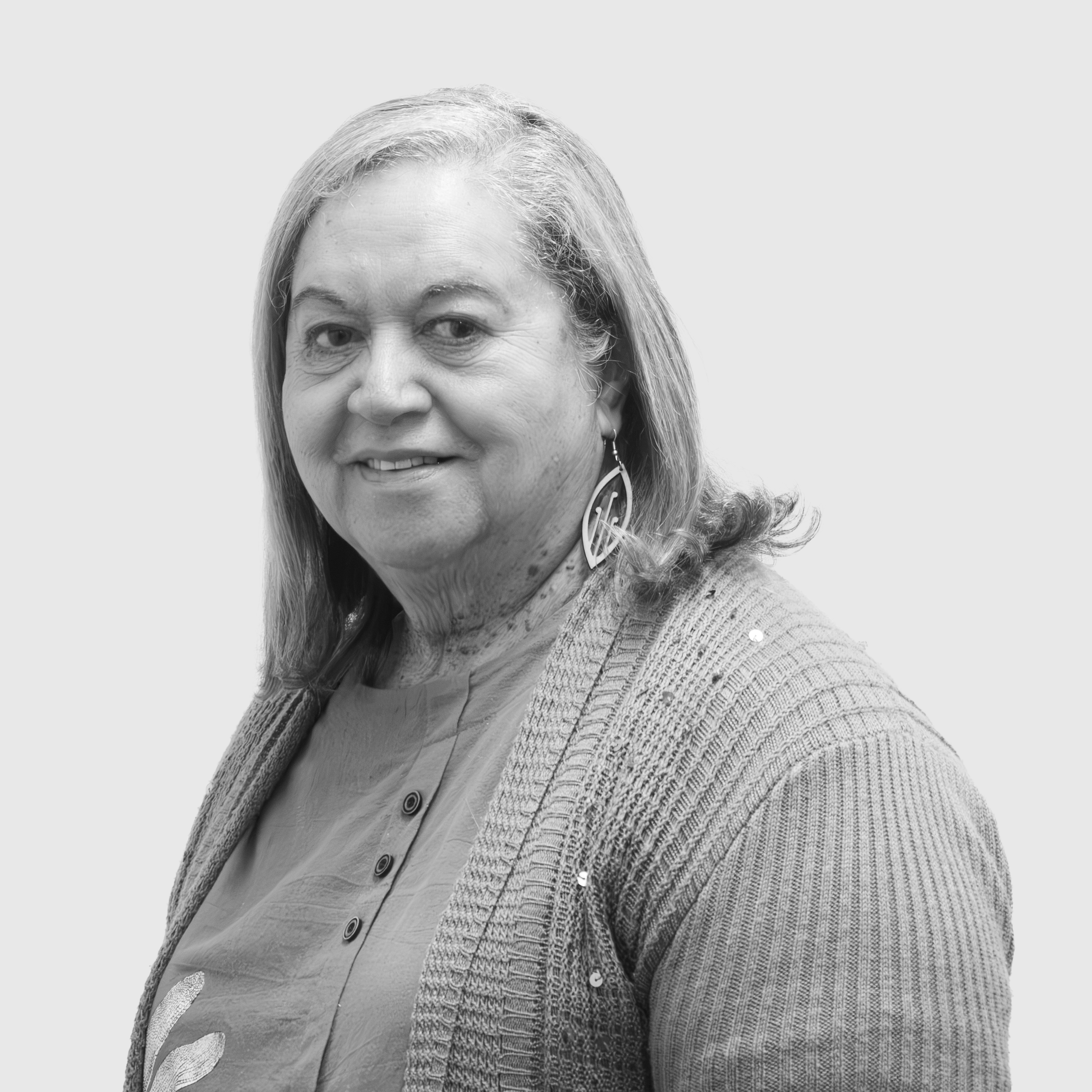 A black and white photo of a smiling middle-aged woman with long hair, wearing earrings, a button-up shirt, and a tweed blazer, against a plain background.