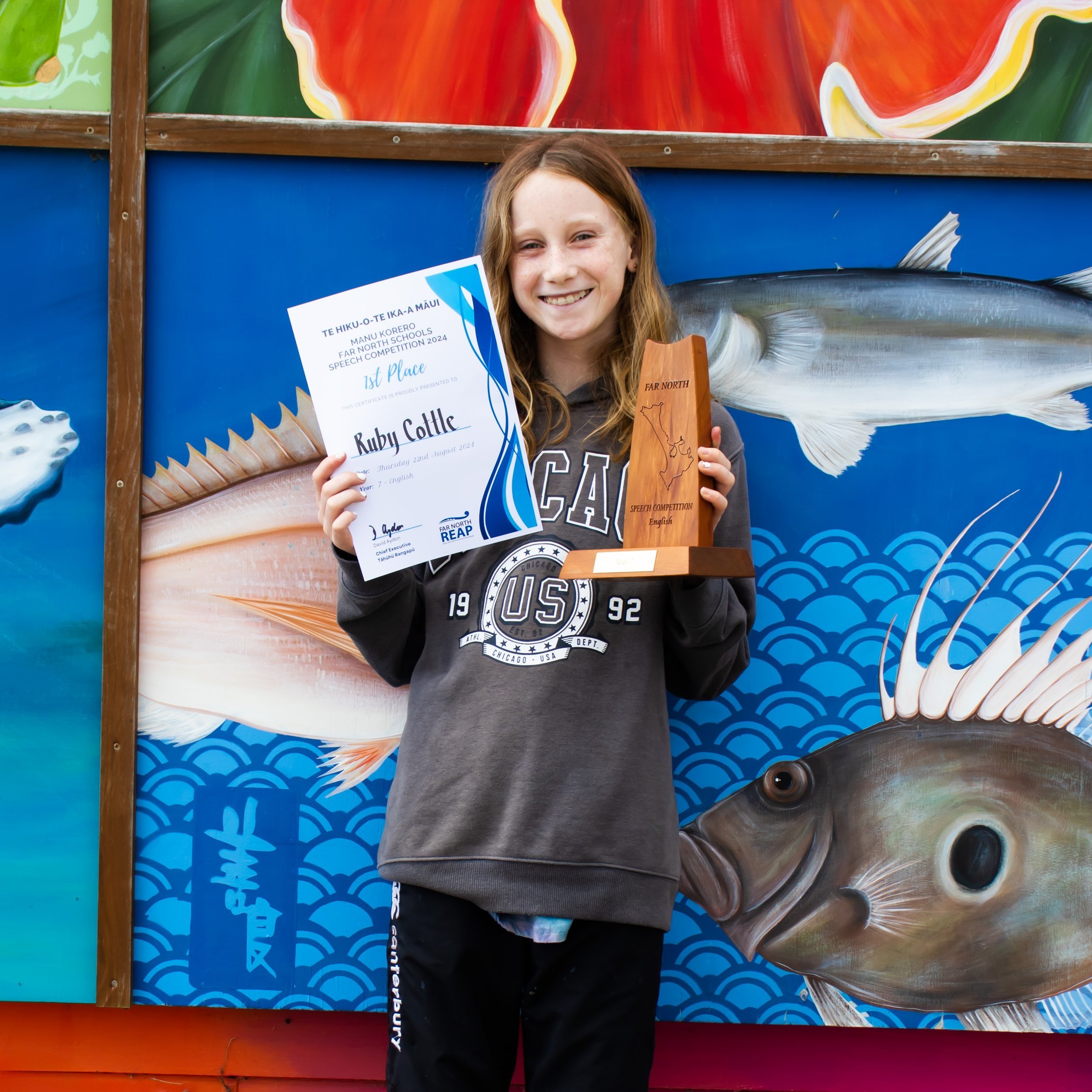 A young girl smiling and holding a speech competition award and a wooden trophy, standing in front of a colorful mural with fish and ocean waves.