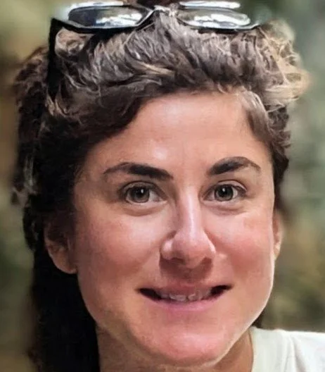 Close-up of a woman with short curly brown hair and sunglasses on her head, smiling outdoors.