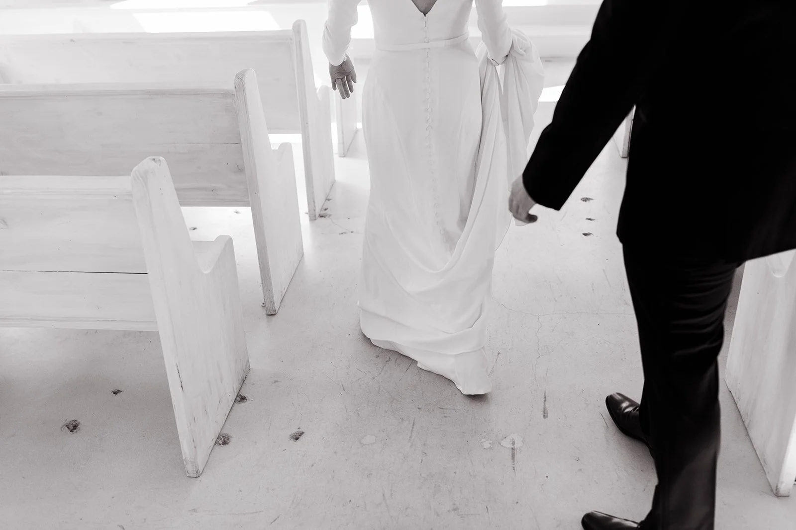 Bride and groom walking down church aisle, bride in white gown holding skirt, groom in black suit, white pews around.