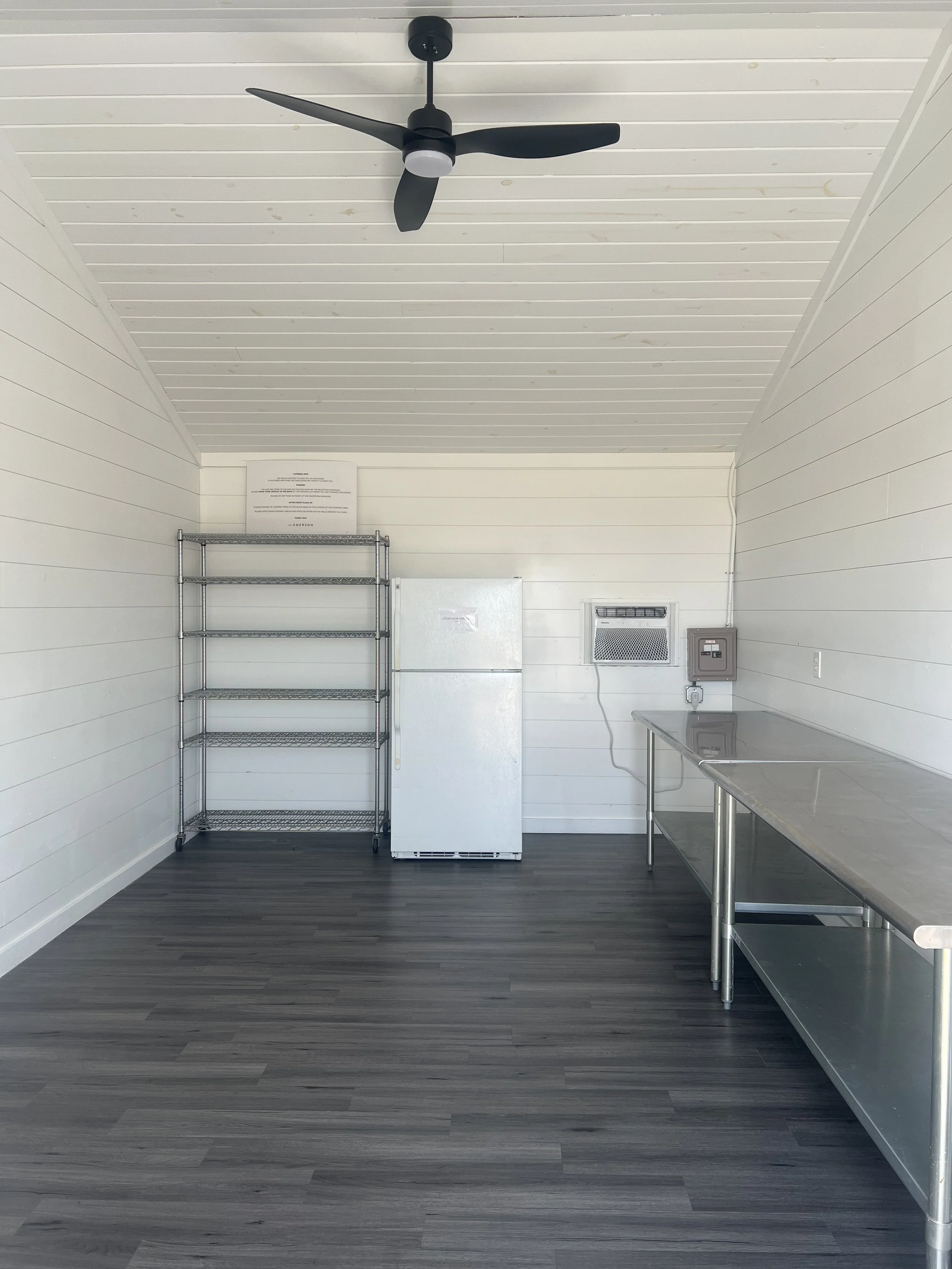 Minimalist room with white shiplap walls, dark wood floor, ceiling fan, metal shelving unit, white refrigerator, and stainless steel counter with sink.