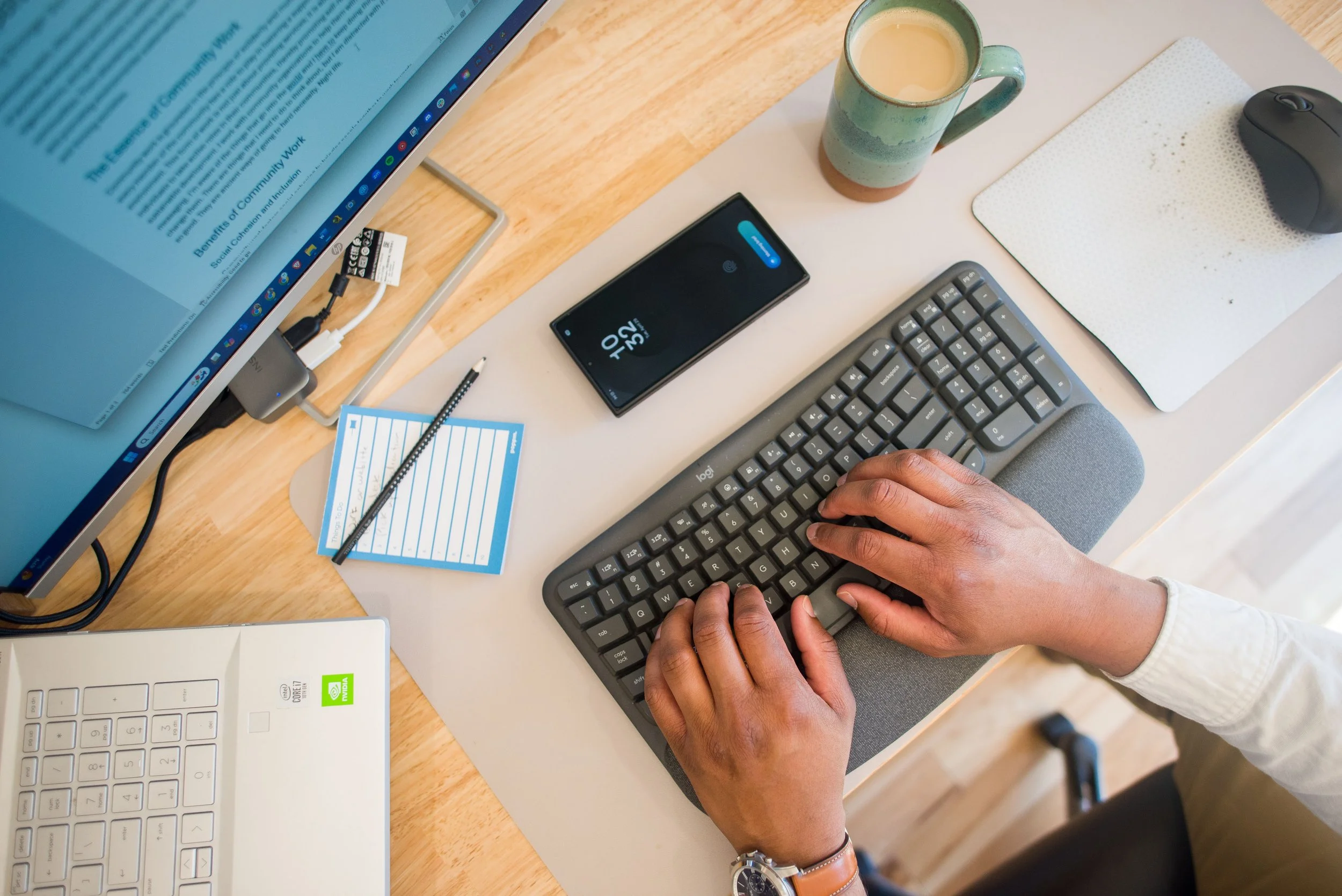 Person typing on a gray keyboard at a workspace with a computer monitor, smartphone, coffee mug, wireless mouse, and a notepad with a pencil.