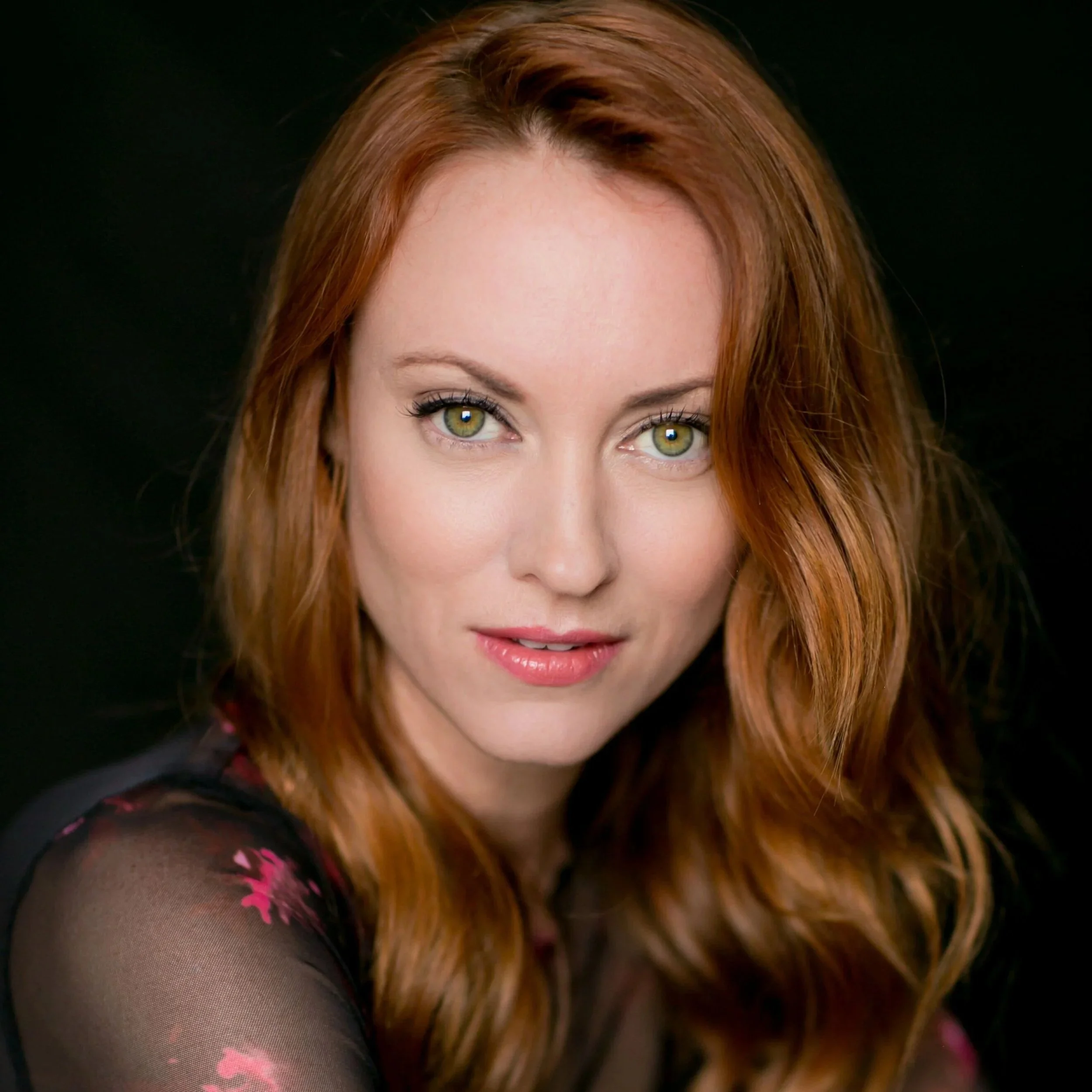 A woman with red hair and green eyes wearing a black top with pink floral patterns, smiling slightly, against a black background.