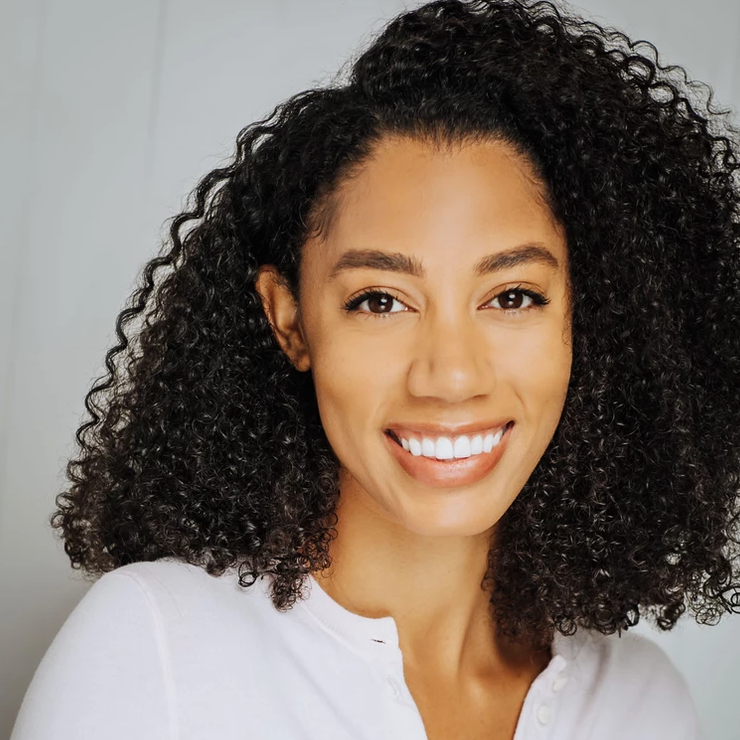 A woman with curly black hair and a broad smile, wearing a white shirt, posing against a light gray background.