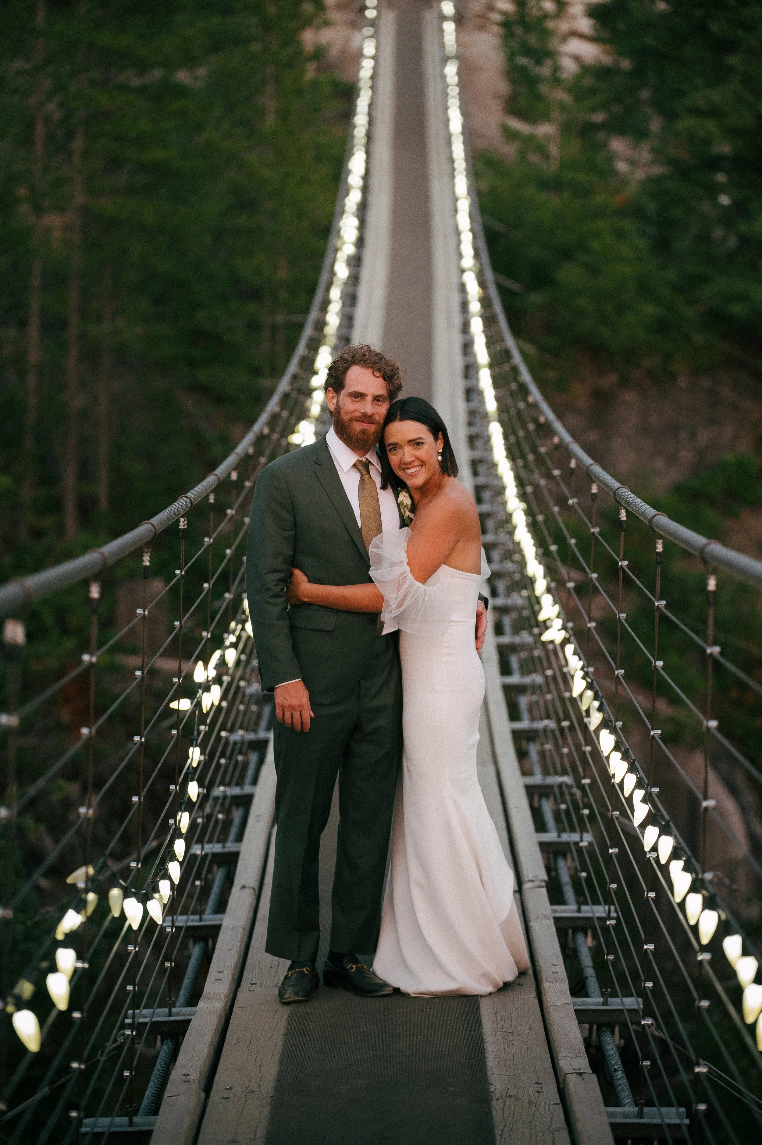 A newlywed couple standing on a suspension bridge with string lights, surrounded by lush green trees, during twilight.